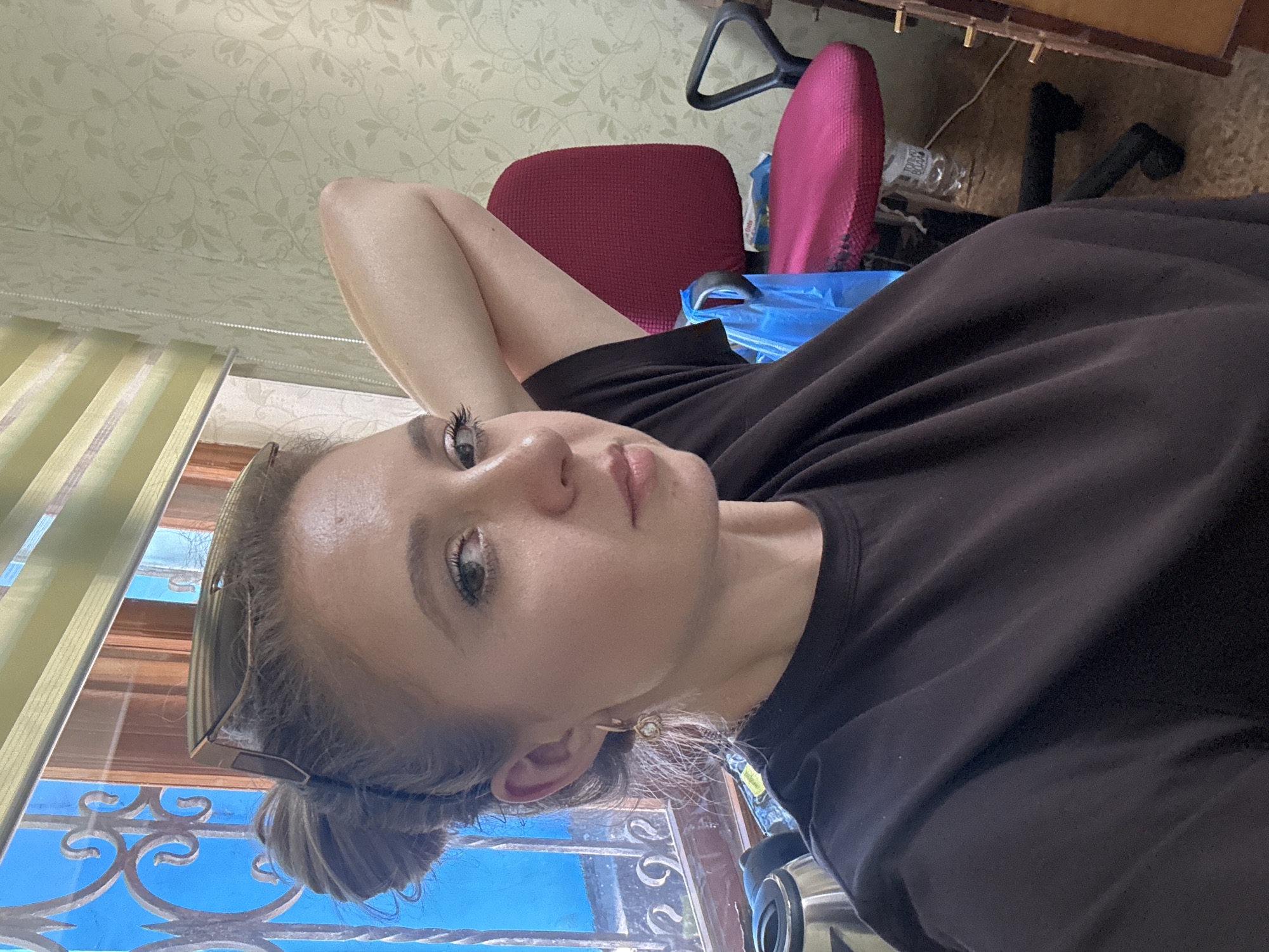 A woman is photographed from the chest up, facing slightly to her right, with her left arm bent and resting behind her head. She has her hair pulled up in a bun and is wearing a dark t-shirt. Sunglasses are perched on top of her head. The background shows a window with blue panes and decorative metalwork, a set of horizontal blinds, and a patterned wallpapered wall. To the right, a pink office chair with black accents is visible. A blue plastic bag and a water bottle are on the floor near the chair. The scene appears to be indoors, likely in a home or office setting.