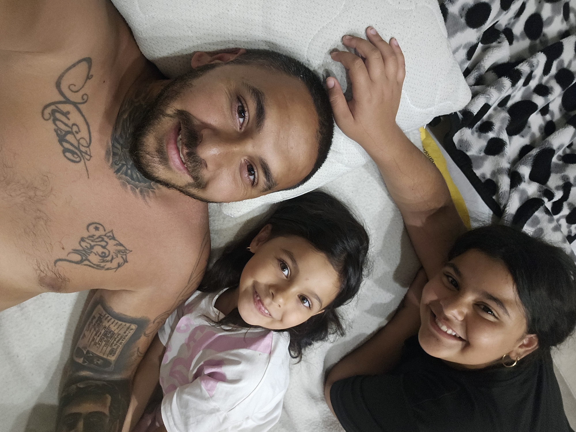 In this media file, three individuals are lying down together, likely on a bed, as indicated by the presence of a pillow and a blanket with a spotted pattern. The setting appears to be indoors, and the lighting suggests it is daytime.
The subjects are two children and an adult male. The adult male is lying on his back, with his chest and face visible. He has several tattoos, including script on his neck and chest, and what appears to be a wolf or similar animal tattoo on his upper arm. He is looking towards the camera with a slight smile. One child, a girl, is lying next to him, also on her back. She has long, dark hair and is wearing a white t-shirt with pink detailing. She is smiling broadly and looking at the camera. Another child, presumably an older girl, is lying on the other side of the adult male, her arm resting on his. She also has dark hair and is wearing a black t-shirt. She has a wide smile and is looking towards the camera. The overall emotion conveyed by the subjects is happiness and closeness.
There is no discernible text in the image, except for the script on the adult male's tattoos, which are not legible. The presence of a spotted blanket and the subjects' relaxed poses suggest a casual, familial moment. The location context of Sofia, Bulgaria, is provided but does not offer specific visual cues within the image itself.