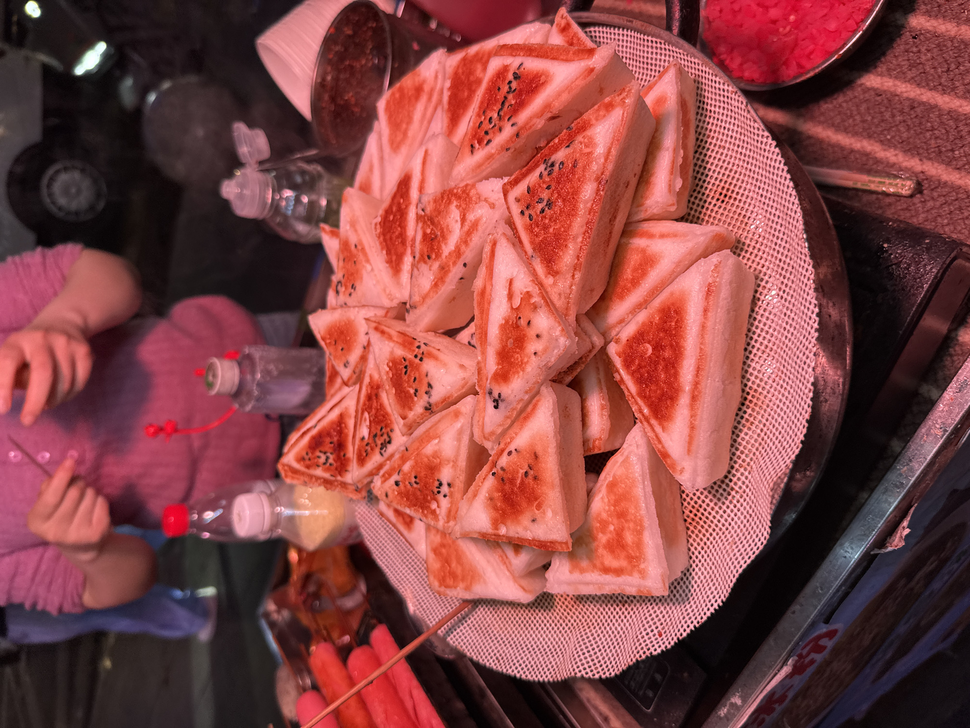 The image depicts a street food scene, likely taken in Yuzhong District, China, judging by the food items and overall atmosphere. A vendor, identifiable by their left arm clad in a pink top, is serving or preparing food. The main subject is a large pile of triangular fried bread or pastries arranged on a mesh-like tray, sprinkled with black seeds (possibly sesame). Condiments or ingredients are visible: a cup of dark sauce, bottles with colored liquids, and a bowl containing a red substance. Red sausages on sticks are also nearby. The overall lighting suggests an evening or nighttime setting.