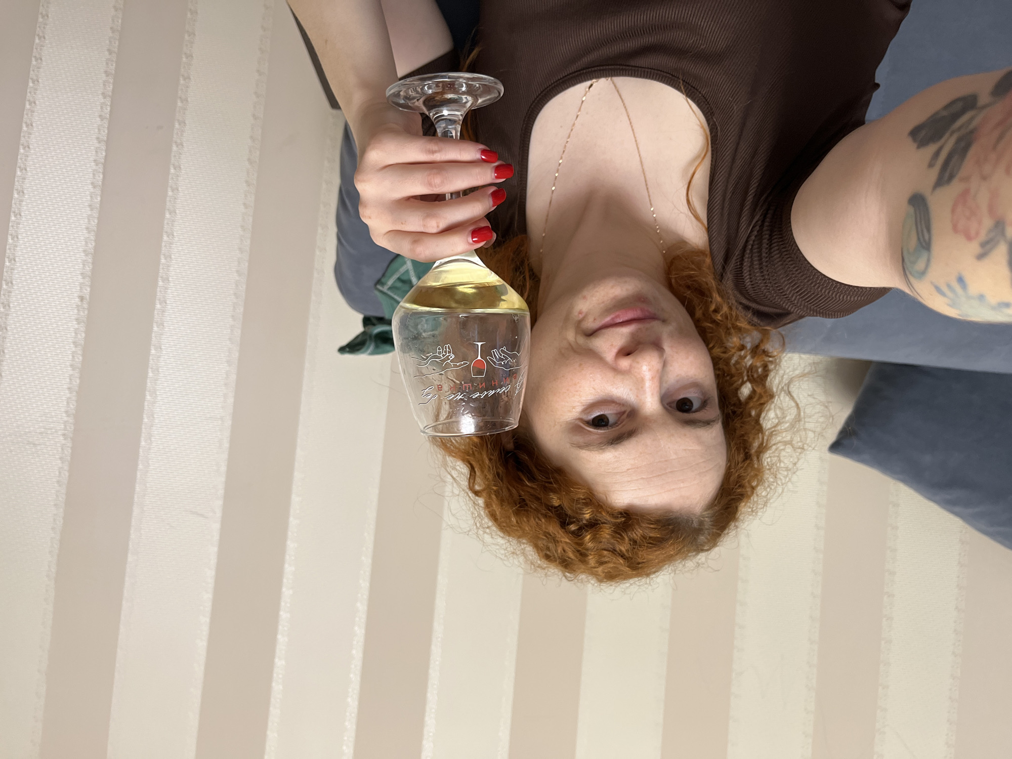 A woman with red curly hair is holding a wine glass filled with a light-colored liquid, likely white wine. She is wearing a brown top and a delicate gold necklace. Her nails are painted a bright red. Her left arm is visible and has a tattoo. The background consists of a striped wall and a blue sofa with pillows. The city of Chaikovsky, Russia is not visible in the image and its location cannot be determined from the information provided.