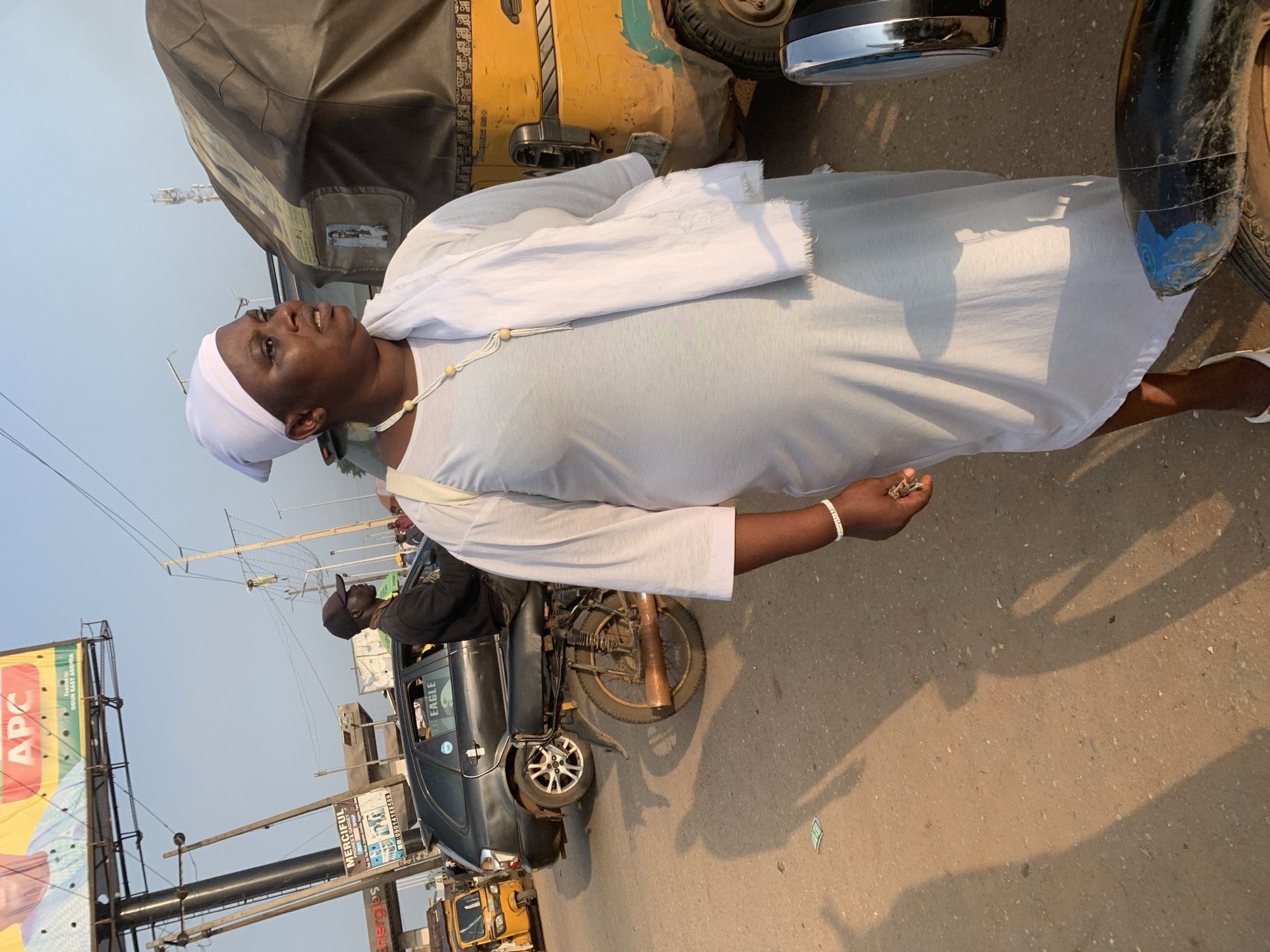 In Shagamu, Nigeria, a woman wearing a white headscarf and a white robe stands on a dusty road next to a yellow auto-rickshaw. She is looking to her right with her mouth slightly open. Around her neck is a beaded necklace, and on her left wrist, she wears a white beaded bracelet. She is holding keys in her right hand. Behind her, a black car with the word "EAGLE" on it is parked. A man wearing a black cap and black shirt is sitting on a motorcycle behind the car. In the background, there is a large billboard advertising "APC" and another billboard with advertisements for "Energis." Power lines and cell towers are visible against the sky, indicating an urban or semi-urban environment. The sun casts long shadows on the road, suggesting it is either early morning or late afternoon.