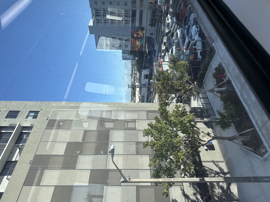 The image displays an urban scene in Hialeah, United States of America, captured from an elevated perspective through a glass window, indicated by prominent reflections. On the left, a tall, multi-story concrete building with upper-level balconies featuring black railings dominates the foreground. Its lower and middle sections are partially enveloped by a light-colored, square-patterned screening material, creating a textured facade. A grey street light on a utility pole is centrally positioned on the left side, extending upwards. Below this, a paved sidewalk area incorporates a landscaped strip containing two young, staked trees and smaller vegetation, along with a visible red fire hydrant.
To the right of the primary building, a large outdoor parking lot is densely filled with various automobiles, including sedans, SUVs, and trucks. Beyond the parking area, another multi-story building, lighter in color with numerous windows and balconies, is present. A vibrant mural, depicting a stylized woman in sunglasses against a tropical sunset with palm trees, adorns a structure behind the parking lot. The background extends to reveal additional urban architecture under a clear, bright blue sky with no discernible clouds. The entire scene is illuminated by direct sunlight, creating distinct shadows on the ground and buildings.