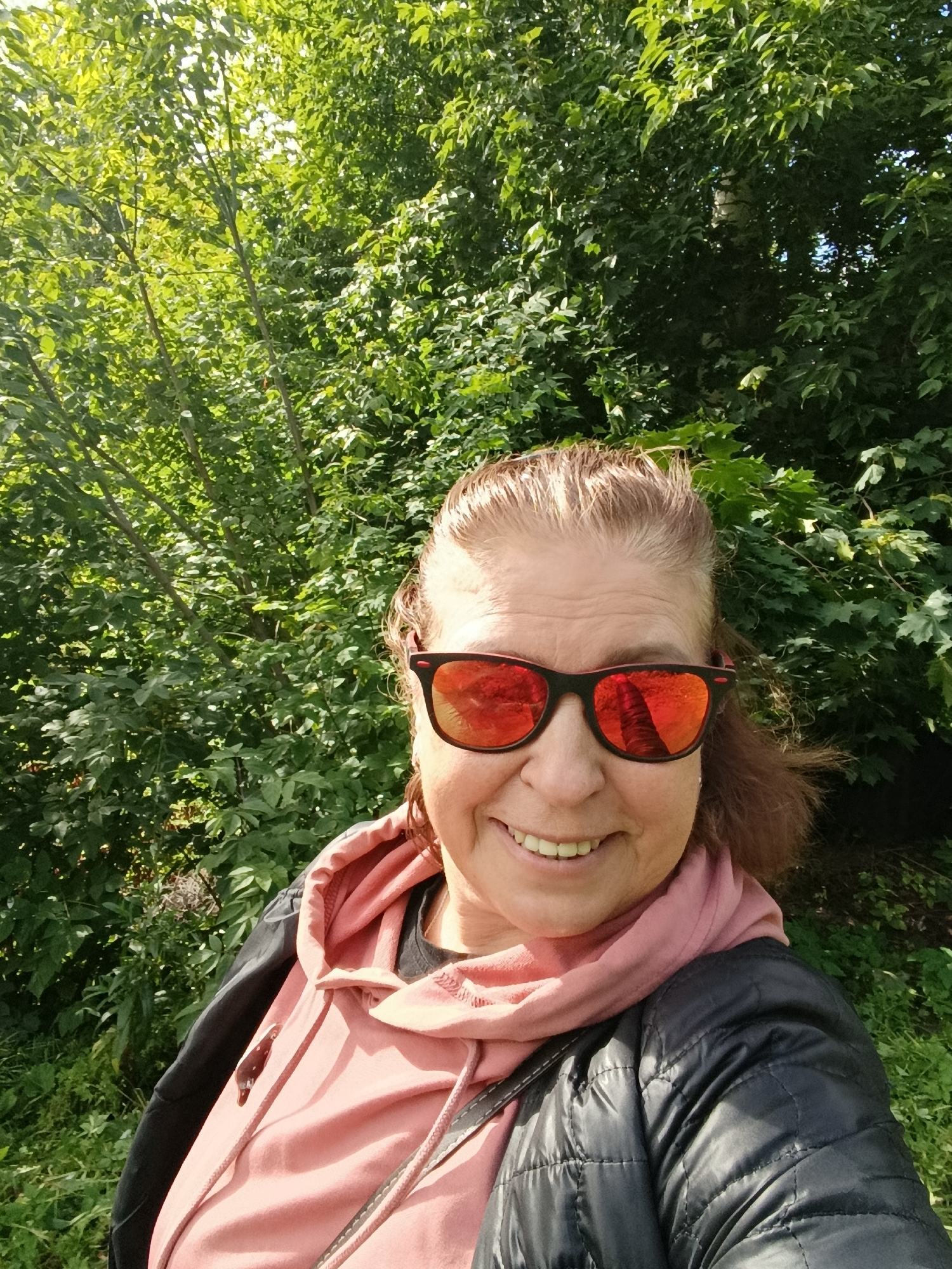 A woman with blonde hair, wearing sunglasses with red lenses and a pink hoodie layered under a black puffer jacket, smiles at the camera. She is outdoors, with a backdrop of lush green trees. The reflection in her sunglasses shows a tall, bare tree trunk. The scene appears to be a casual, sunny day in a natural setting, possibly a park or wooded area.