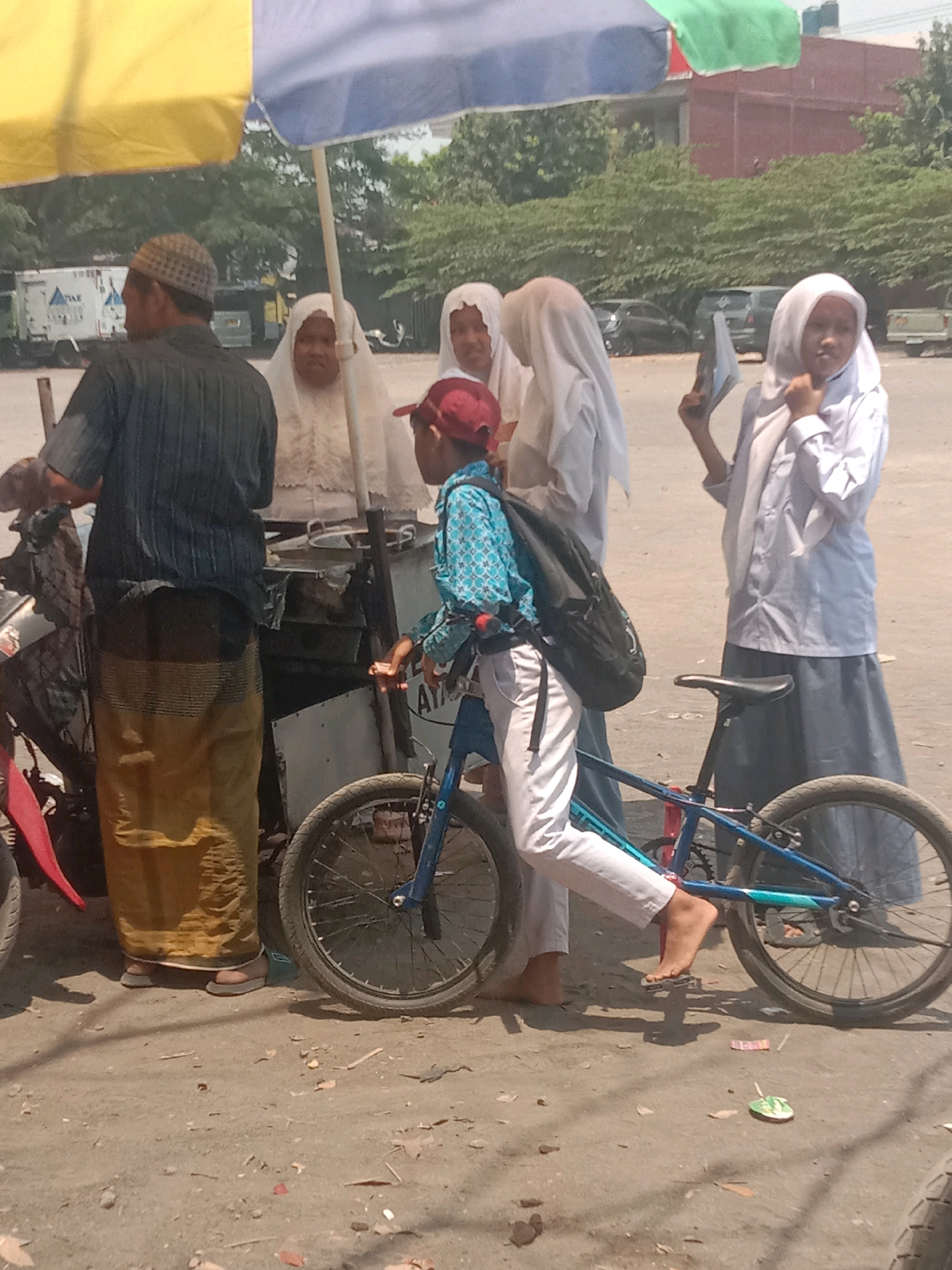 A group of people are gathered under a large, multi-colored umbrella, likely at a street market or gathering in Ngempit, Indonesia. In the foreground, a young boy wearing a blue patterned shirt, a red baseball cap, and white pants is standing next to a blue bicycle, appearing to be preparing to ride it or adjust it. He has a black backpack slung over his shoulder. To his left, a man in a dark striped shirt and a yellow sarong stands facing away from the camera, interacting with a food stall or cart. Behind him, three individuals are visible, two women wearing white hijabs and one possibly a child also in white. The woman in the middle appears to be looking at the man. Another woman, wearing a white hijab and a light blue uniform, is standing further to the right, holding a document or book. The environment is an unpaved, dusty outdoor area with trees and buildings visible in the background. Several vehicles, including a truck and cars, are parked in the distance, suggesting a roadside location within the city.