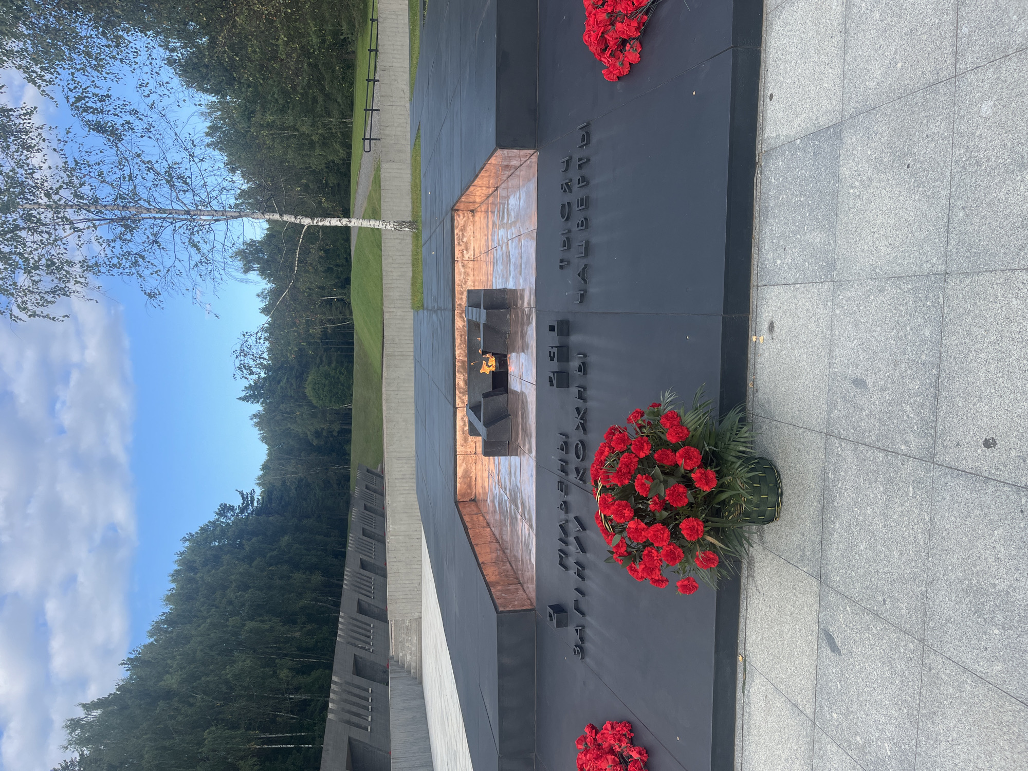 The image captures a somber memorial site, likely a place of remembrance, situated in the Каменскі сельскі Савет, Belarus. The primary subject is a stark, dark monument featuring raised, blocky elements within a rectangular pit that appears to be filled with reflective copper or bronze material. A flame burns in the center of this pit, casting a warm glow. Surrounding the pit are flat, dark surfaces inscribed with what appear to be Cyrillic letters. Red carnations are placed as tributes on the monument's surface, with one arrangement in a woven green basket situated prominently. In the background, a line of tall, narrow concrete structures with angled openings stretches into the distance, set against a backdrop of dense green trees and a clear blue sky with scattered clouds. A solitary, slender birch tree stands to the right of the central monument, its white bark and sparse foliage contrasting with the darker elements of the memorial. The ground in the foreground is paved with large, grey granite tiles. The overall scene conveys a sense of solemnity and remembrance.