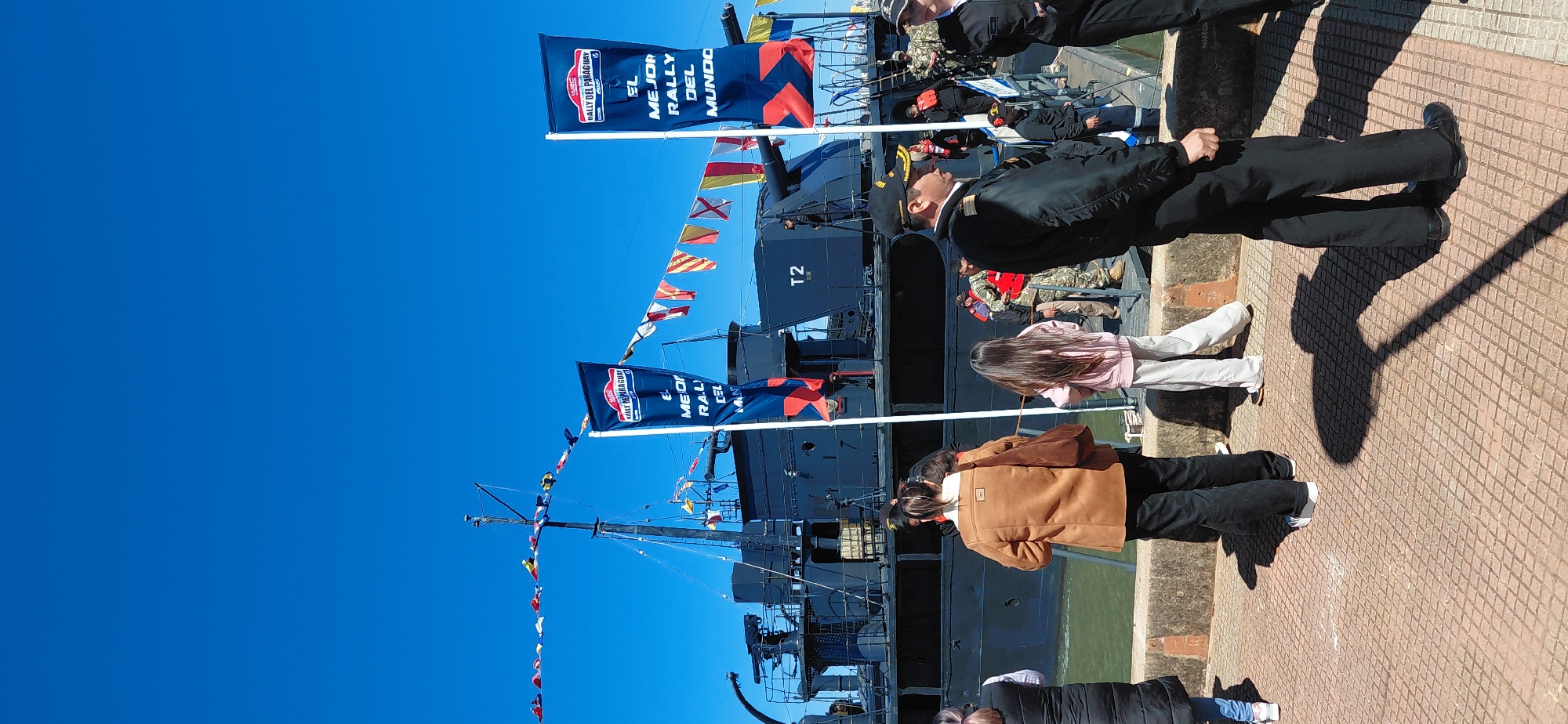 The image depicts a maritime event in Encarnación, Paraguay, likely a festival or exhibition involving naval vessels. A dark-colored ship, possibly a historical or military one, is docked and prominently features two large blue banners with white text, one reading "RALLY DEL PARAGUAY" and another stating "EL MEJOR RALLY DEL MUNDO". The ship's deck is visible, with what appear to be cannons and other military equipment. Numerous signal flags are strung across the mast of the ship and between structures, adding a festive atmosphere.
A group of people, some in casual attire and others in what appear to be naval uniforms, are gathered on a paved walkway next to the dock. In the foreground, a person in a dark uniform with a cap is facing to the right, with their profile visible. Another person in a similar uniform is seen to the right of the frame. Towards the left, a woman in a brown jacket and dark pants is standing with her back to the camera, and a young girl in a pink top and light pants is next to her. Other individuals are seen in the background, some wearing life vests, suggesting a possible maritime safety demonstration or a tour of the ship. The sky is a clear, bright blue, indicating a sunny day. The walkway has a patterned tile surface with diamond shapes. The overall scene suggests a public event that is drawing a crowd to view the naval vessel.