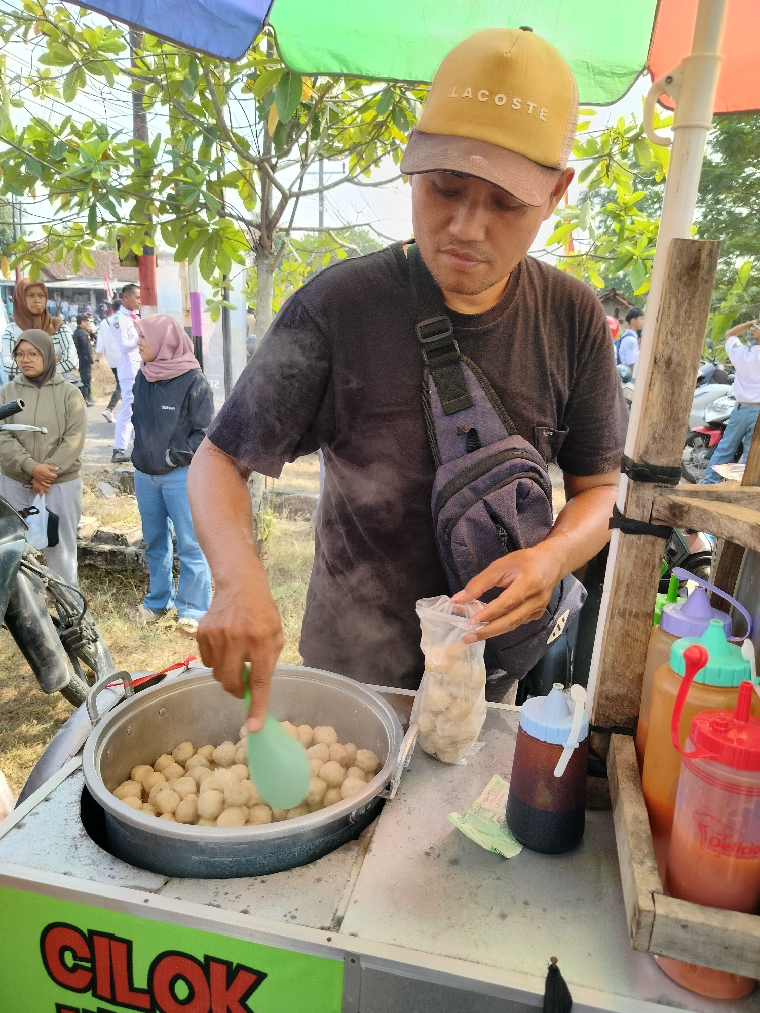 The image shows a man wearing a yellow "LACOSTE" baseball cap and a black t-shirt, serving food from a cart. He is using a green ladle to scoop food from a large metal pot filled with small, round meatballs. The cart has a sign that says "CILOK" in large green letters. To the right of the cart are several condiment bottles, including one with a red cap containing a red sauce and another with a purple cap and a blue lid containing a dark liquid. Behind the man, several people are visible, including a woman wearing a brown hijab and a light green hoodie, and another woman in a pink hijab and a black jacket. In the background, there are trees, a building, and what appears to be motorcycles. The scene appears to be outdoors, possibly on a street or market. The overall impression is a street food vendor in action, likely in Indonesia, given the "CILOK" sign which refers to a popular Indonesian snack.