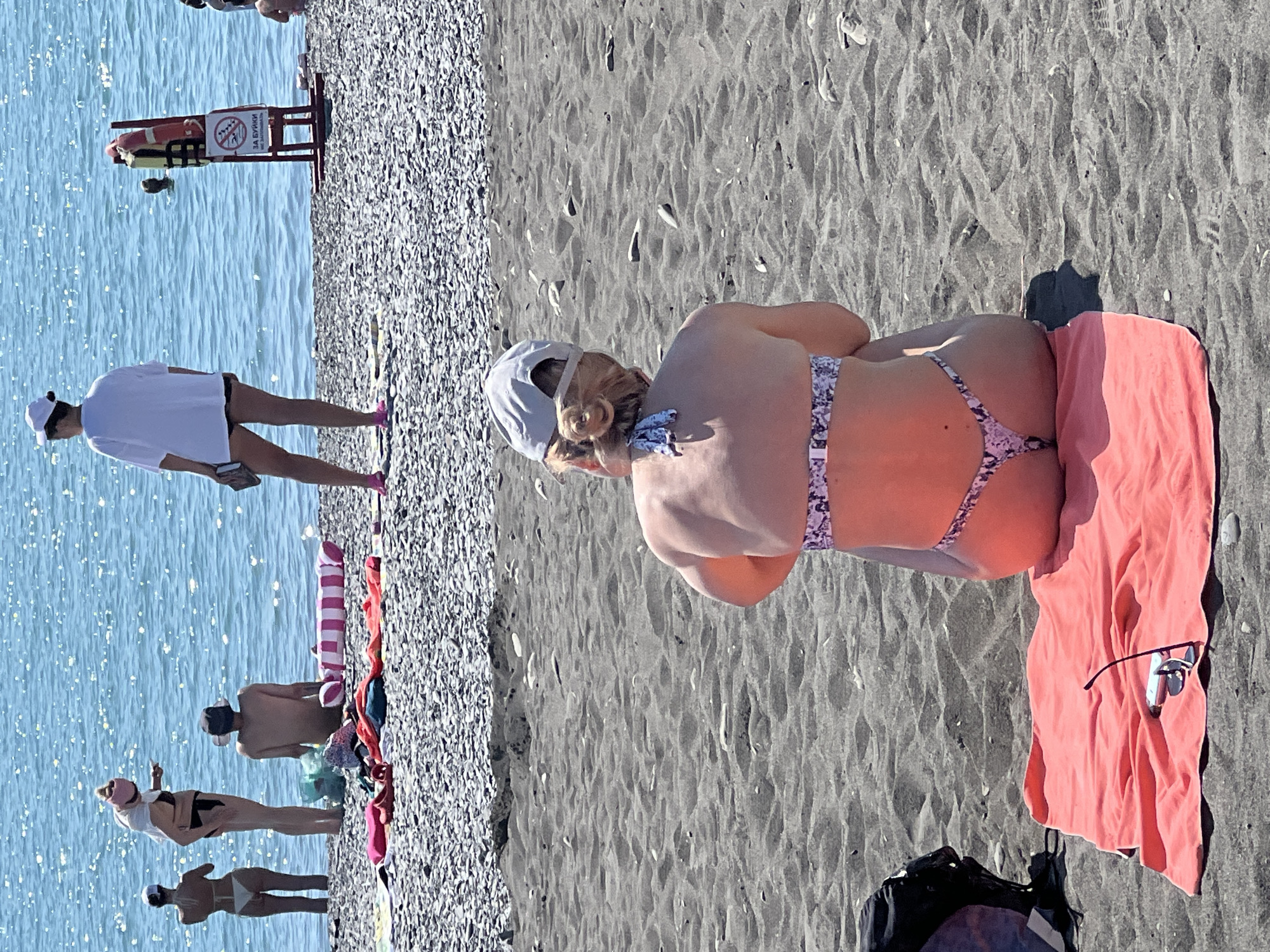 A woman wearing a white baseball cap and a patterned bikini is sitting on a bright orange towel on a black sand beach. She is facing away from the camera. Her hair is pulled back in a bun. Next to her on the towel is a pair of sunglasses. To her left, a black backpack is partially visible. In the background, the blue ocean sparkles under the sunlight. Several other people are visible on the beach, some in the water and others on the shore, along with beach towels and a life preserver on a stand. The scene appears to be at a beach in Sochi, Russia.