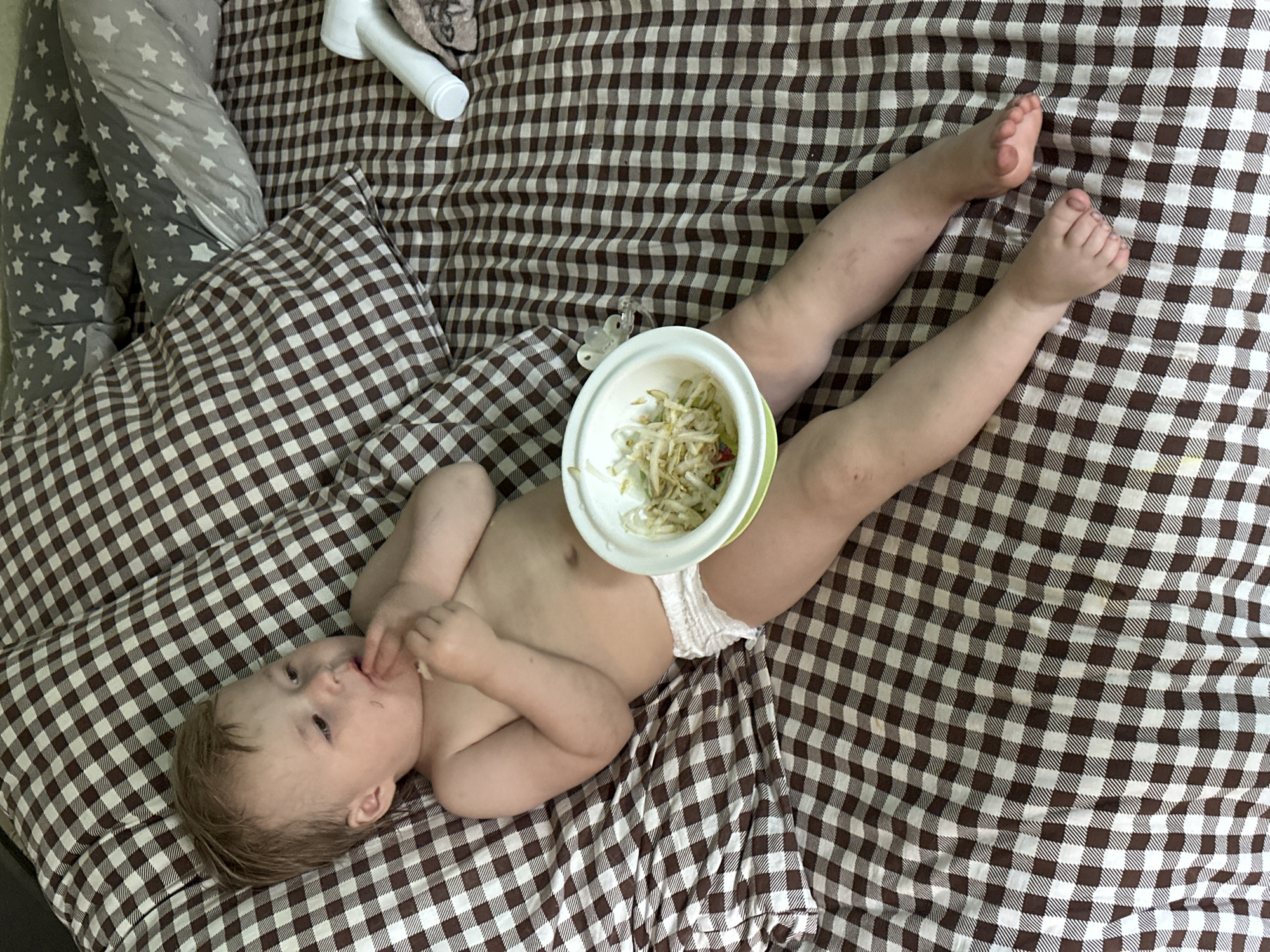 A baby in a diaper lies on a brown and white checkered bedspread. A bowl of bean sprouts sits on the baby's stomach. The baby is lying on its back with its legs stretched out and is reaching for its mouth with its right hand. To the left of the baby is a grey pillow with white stars. Near the top left corner of the image, next to the grey pillow, is a white bottle and a piece of brown fabric. A clear pacifier is lying next to the baby's leg, close to the bowl of bean sprouts. The location is not specified, so Nizhny Novgorod, Russia, cannot be confirmed.