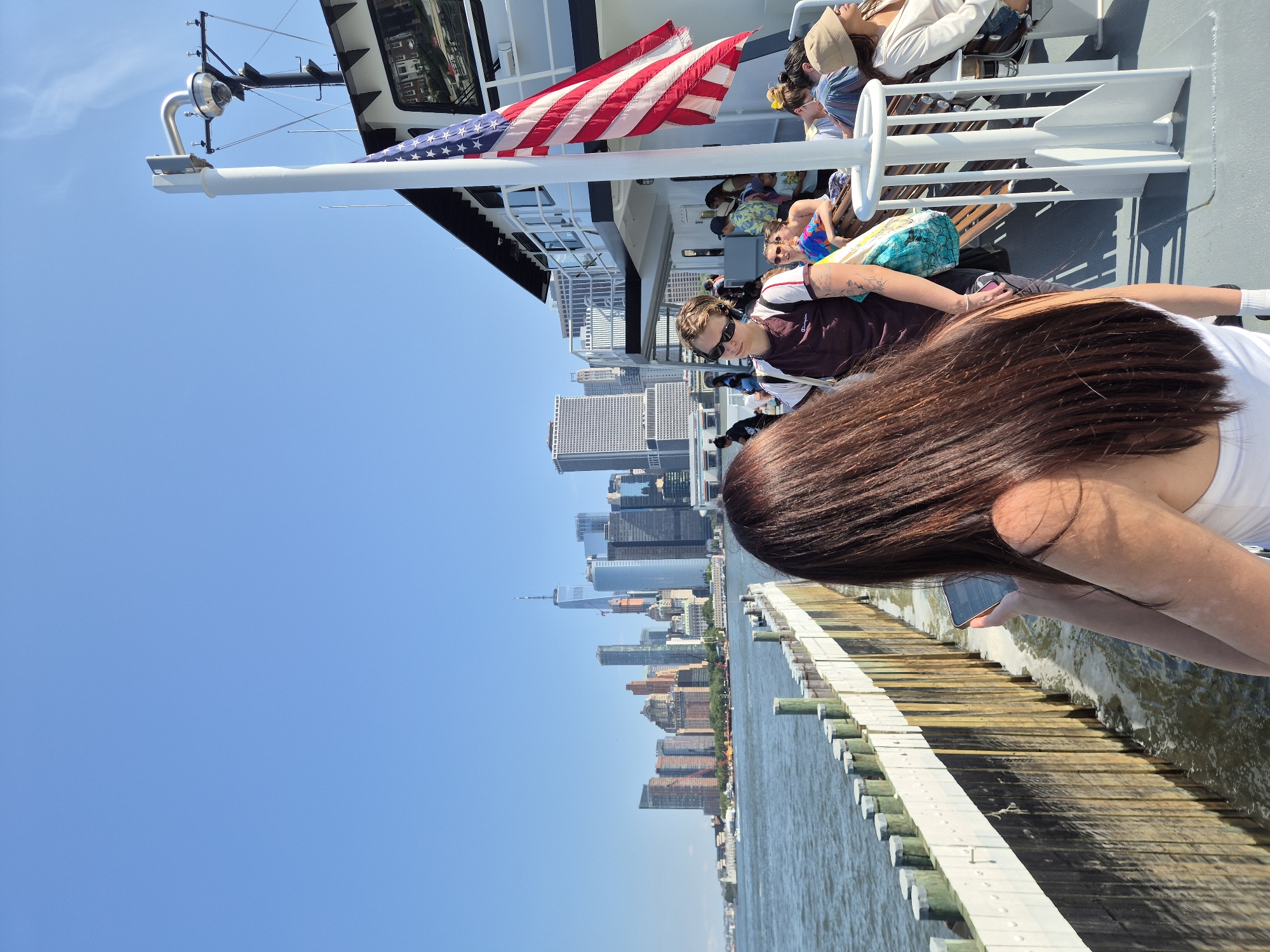 An outdoor, low-angle medium shot captures a group of people on a ferry with the New York City skyline in the background. The ferry deck is grey, with a wooden railing along the side. The water is choppy and blue. In the foreground, a woman with long, dark brown hair is looking down at her phone. She is wearing a white top. To her left, a person wearing a maroon t-shirt and sunglasses is looking forward. They have tattoos on their arm. Behind them, other passengers are visible, some sitting on wooden benches. An American flag is attached to a pole on the ferry. The skyline, featuring numerous skyscrapers, is visible across the water under a clear blue sky.