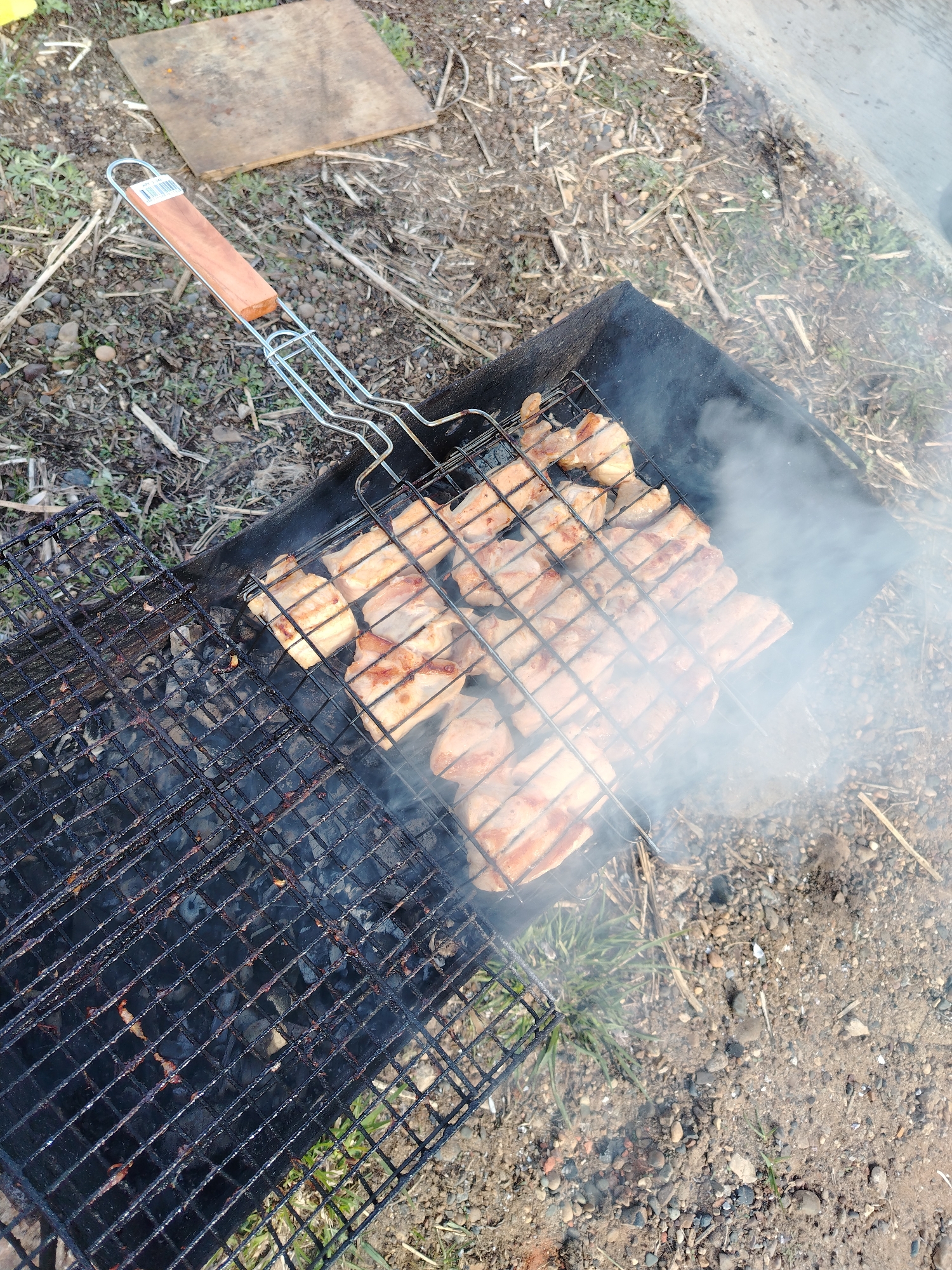 An overhead, slightly angled shot captures an outdoor grilling scene, where pieces of meat are cooking on a metal grill grate over hot charcoal in a black brazier. The meat, likely pork or chicken, is browning with visible grill marks, indicating it is actively cooking. A significant amount of white smoke billows upwards from the grill, partially obscuring the food on the right side of the frame.
The grill grate features a wooden handle, on which a small, unreadable product label with a barcode is visible. Beneath the cooking meat, glowing charcoal can be seen through the grate, providing the heat for the grilling.
The setting is an informal outdoor area with light brown soil, scattered pebbles, and patches of dry grass and green weeds. A weathered, rectangular wooden board lies on the ground in the upper left section of the image. Along the upper right edge, a concrete surface or path is visible. The scene is brightly lit, suggesting it is daytime. No people are visible. The activity taking place is outdoor barbecue, characteristic of a leisure event in a location like Chaikovsky, Russia.