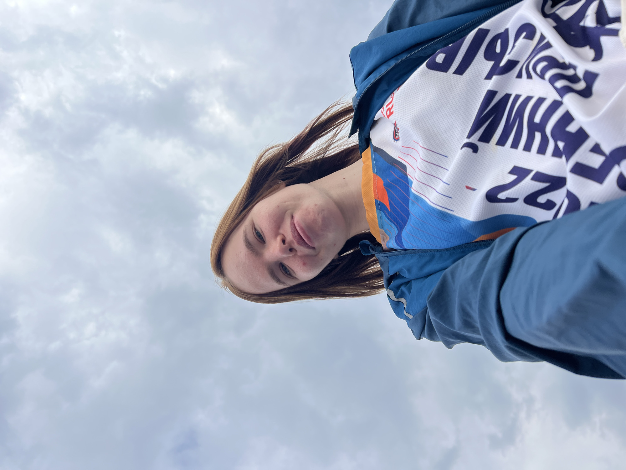 A woman with auburn hair is taking a selfie outdoors under a cloudy sky. She is wearing a blue jacket and a white shirt with blue and orange accents and Russian text. The camera angle is from below, looking up at the woman. The background consists of gray, overcast skies. The location is presumed to be Verkh-Sosnova, Russia.