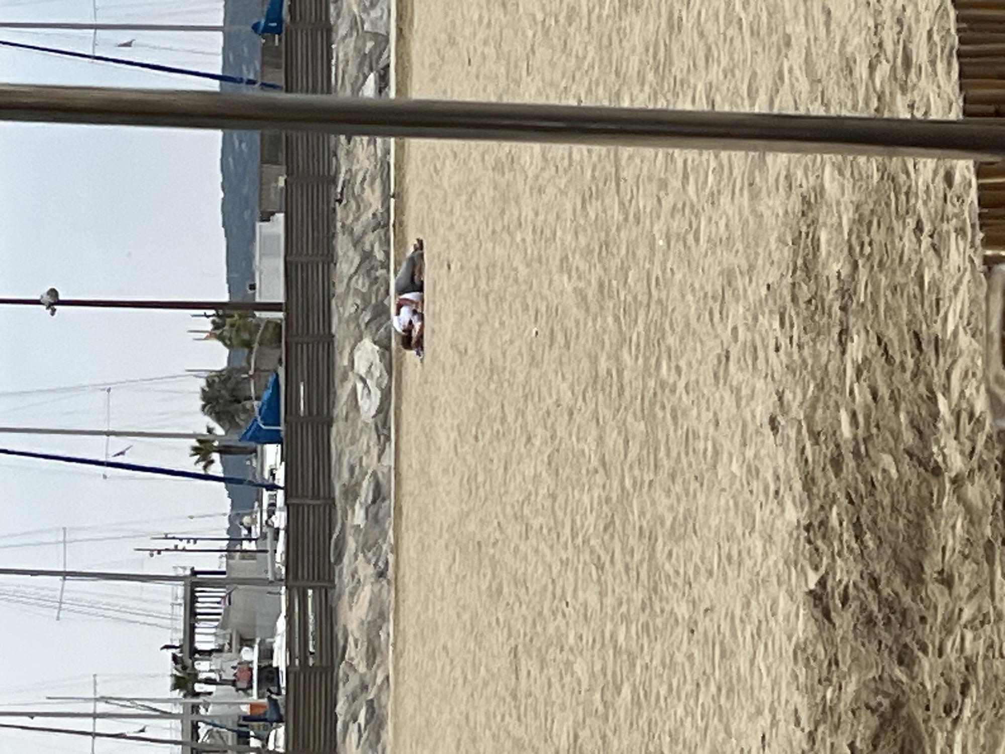 The image depicts a beach in Sainte-Maxime, France. The beach is sandy and there is a person lying on it. In the background, there is a harbor with several sailboats and some buildings. The foreground includes a metal structure.