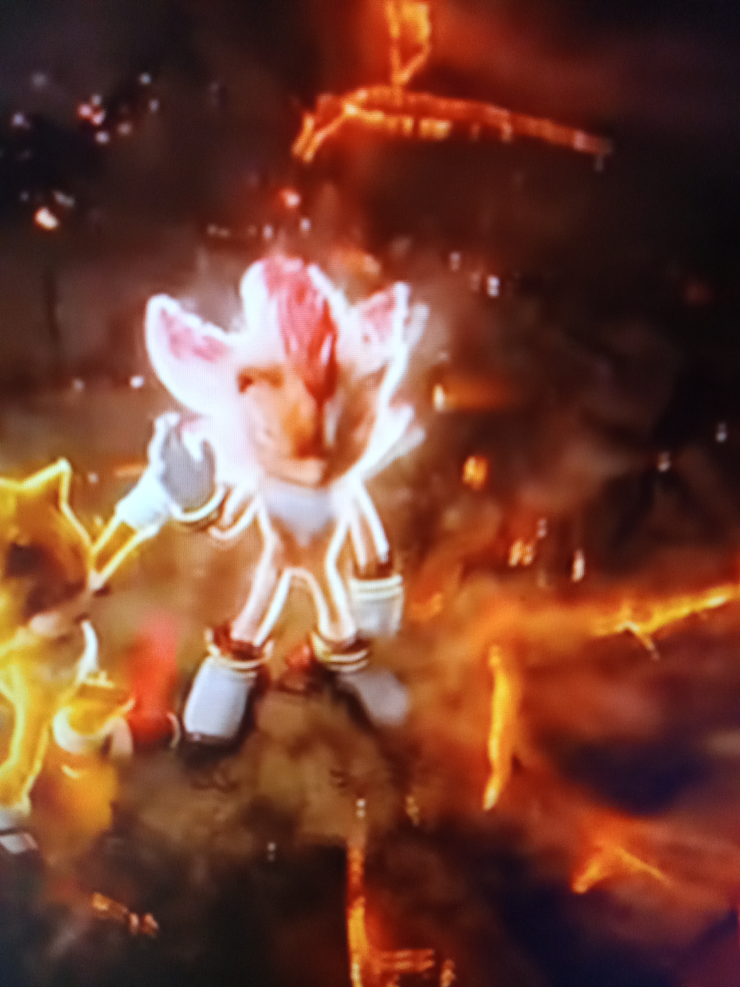 The image is a blurry, digitally captured photograph of what appears to be characters from a video game or animation. In the foreground, a light-colored, humanoid figure with large ears and red accents on its head stands prominently. This figure is emitting a bright, white and pinkish glow, suggesting an energy or transformation state. Its limbs are extended, and it wears white and red accents on its feet and gloves. To the left of this central figure, another character, primarily yellow and outlined in light, is partially visible, appearing to be smaller and potentially reaching out to the central figure. The background is dark and filled with abstract, fiery orange and red patterns, implying an intense or chaotic environment. There are also small, bright white specks scattered throughout the image, possibly representing sparks or debris. The image lacks any discernible connection to the city of Рощин ауылдық округі, Kazakhstan, as it depicts a fictional scene with no real-world geographical indicators.