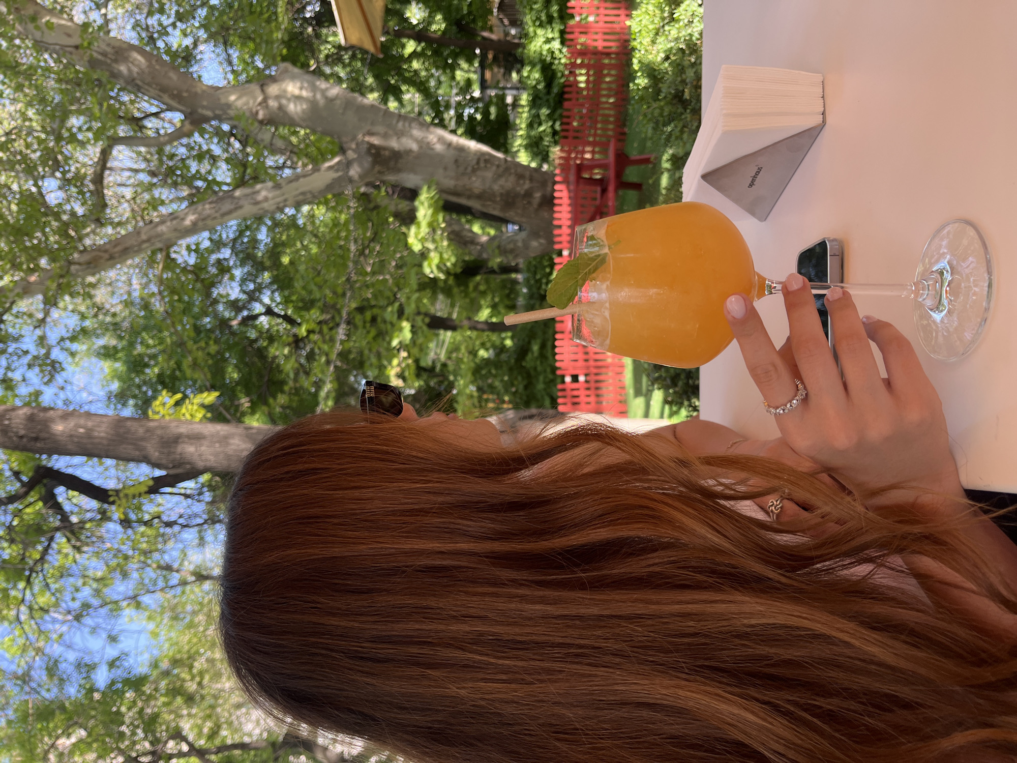 A woman sits at a white table outdoors, holding a glass of orange liquid with ice and a mint garnish. The glass has a straw, and her hand is adorned with a diamond ring. A small, rectangular lighter rests next to her hand. To the left of the glass is a metal napkin holder containing white napkins. Behind the table is a red fence, and beyond that, trees and foliage fill the background. Based on visual cues and the location, the scene likely depicts a cafe or restaurant setting in Yerevan, Armenia.