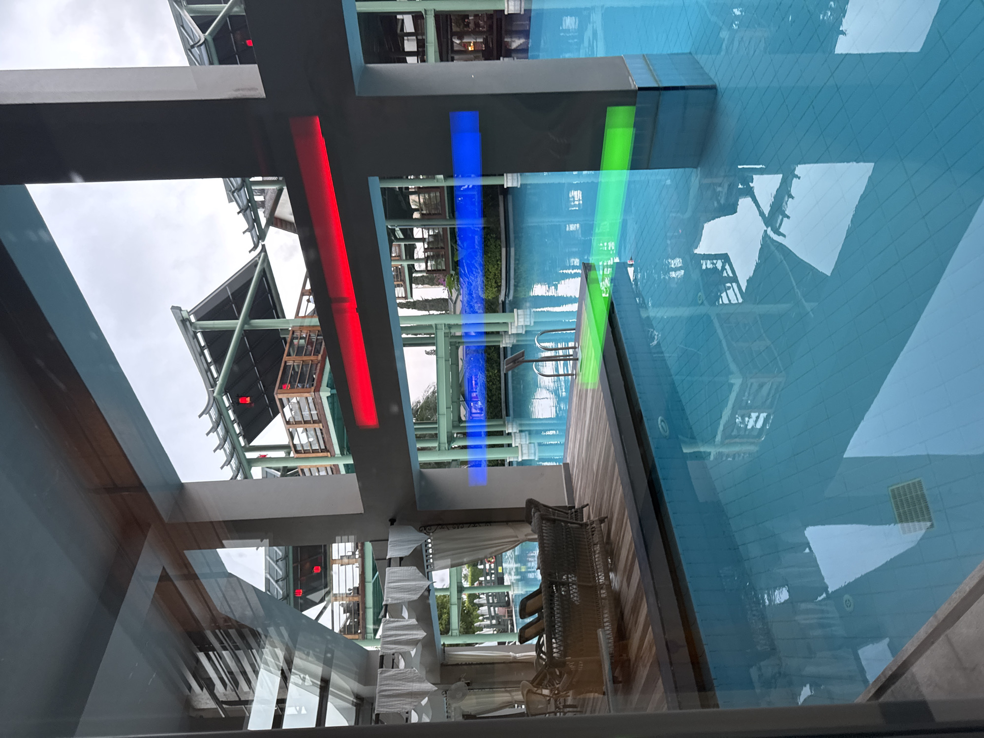 The image shows an indoor swimming pool area with a clear blue water surface. There are wooden decks surrounding parts of the pool, with wicker furniture and white curtains visible on one side, suggesting a lounge or seating area. The architecture features modern design elements, including large concrete pillars and overhead structures with pagoda-like roofs. Colored LED lights are visible, with red, blue, and green strips illuminating sections of the scene. Reflections of the surrounding structures and the sky are visible on the water. The location is identified as Aksu, Turkey, likely a resort or hotel given the amenities. The scene appears to be empty of people.
