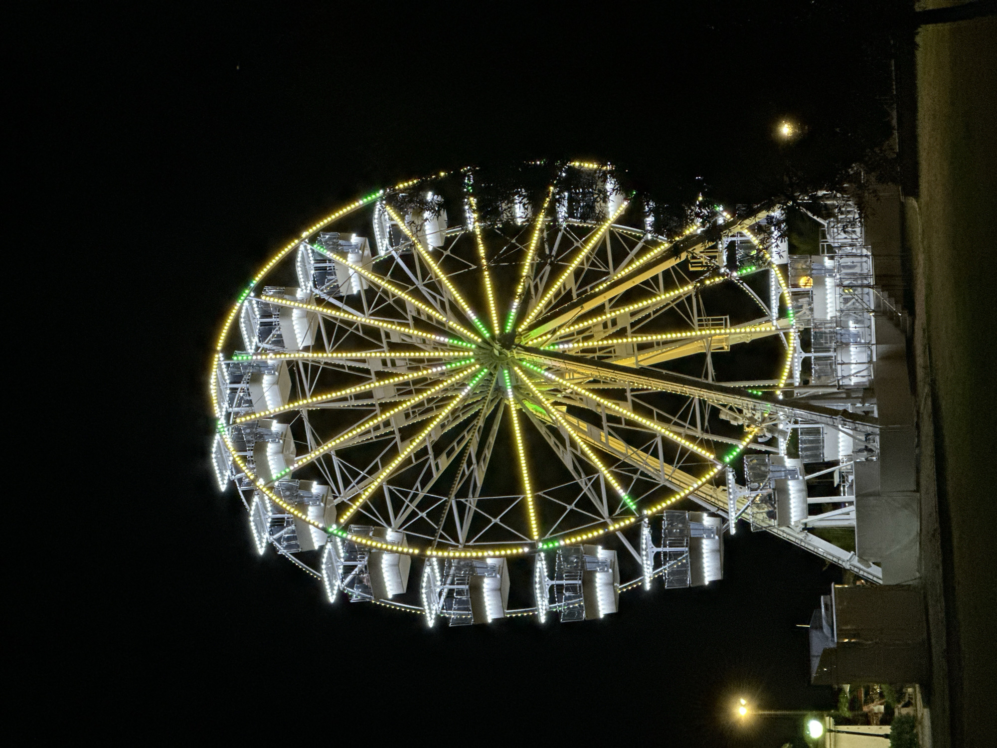 An illuminated Ferris wheel stands prominently against a dark night sky. The wheel is adorned with rows of yellow and green lights that trace its circular structure and spokes. Several white gondolas, each with glass railings and a roof, are attached to the rim of the wheel. Some of these gondolas appear to have people inside.
At the base of the Ferris wheel, a white platform with stairs and railings is visible, suggesting an entrance or waiting area. To the left of the Ferris wheel, a lamppost with a yellow light illuminates the foreground, and a low, rectangular structure is situated near the base of the wheel. The ground in front of the Ferris wheel is covered in green grass. To the right, a dark silhouette of a tree partially obscures the Ferris wheel.
The image was captured at night, likely in an outdoor amusement park or recreational area. The style of the Ferris wheel and its lighting suggest a festive or carnival atmosphere. It's highly probable that this Ferris wheel is located at an amusement park on the Budva Riviera, a popular tourist destination in Montenegro known for its lively atmosphere and entertainment options.