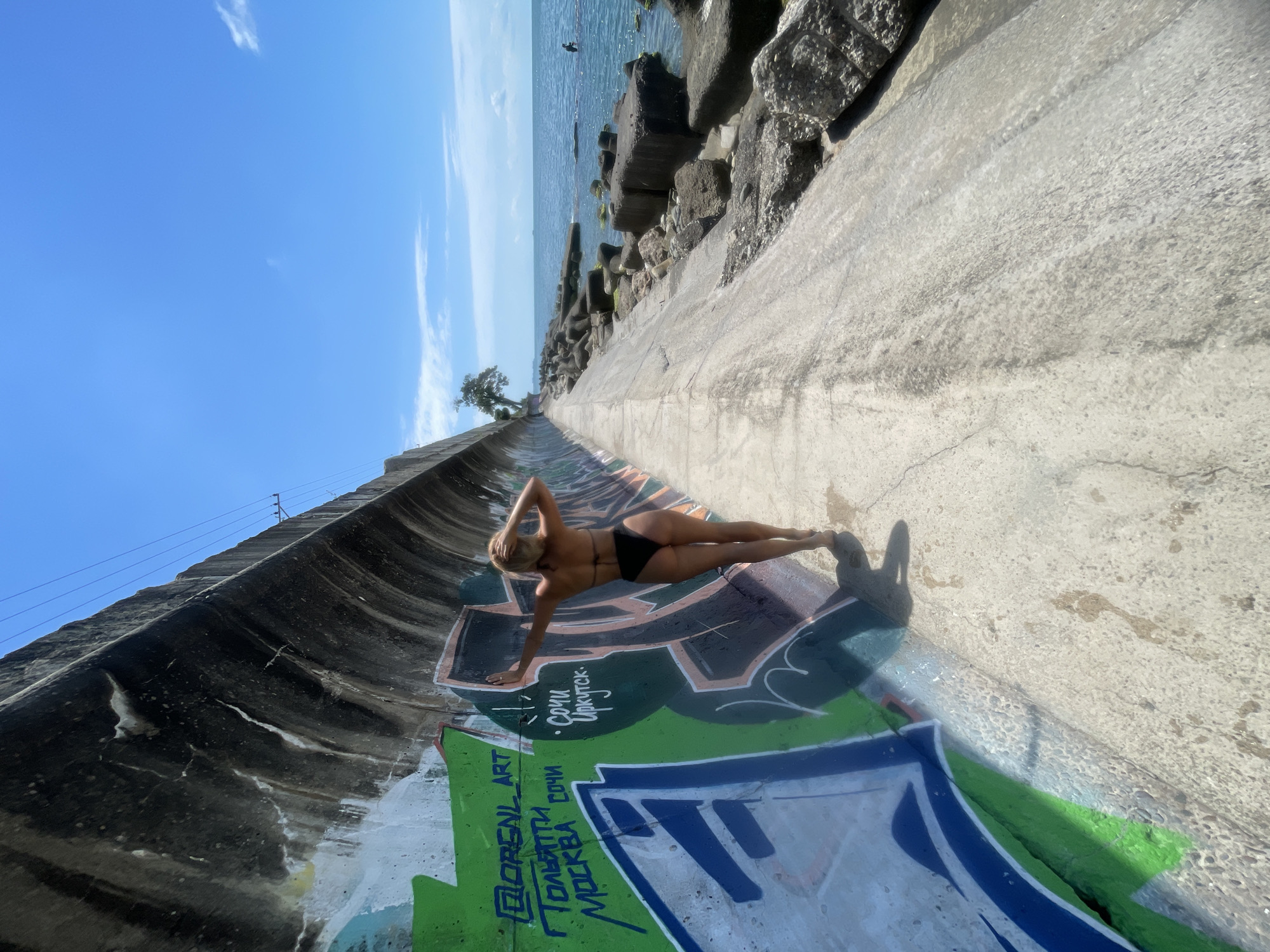A woman in a black bikini is lying on her side on a concrete surface that is covered in graffiti. She is on an upward-sloping path, with her head towards the top of the image and her feet towards the bottom. Her left arm is extended, touching the ground, and her right arm is bent at the elbow with her hand resting on her head. The concrete path is wet in places and appears to be part of a seawall or drainage channel, as the ocean is visible in the background.
The water is a deep blue, with some choppy waves. Large rocks and concrete blocks form a barrier along the edge of the water. A single tree is visible on the left side of the image, growing from the side of the concrete structure. The sky is bright blue with a few wispy white clouds. The overall impression is a sunny day, likely in the late morning or early afternoon, given the strong shadows.
Graffiti covers the lower portion of the concrete path, featuring various colors and styles of spray paint. Some of the visible text is in Russian, including "СОЧИ" (Sochi), "ИРКУТСК" (Irkutsk), "ТОЛЬЯТТИ" (Tolyatti), and "МОСКВА" (Moscow). There are also phrases like "DORENL ART" and "СОЧИ". The woman appears relaxed and is posing for the camera. The setting suggests an outdoor urban or coastal environment.