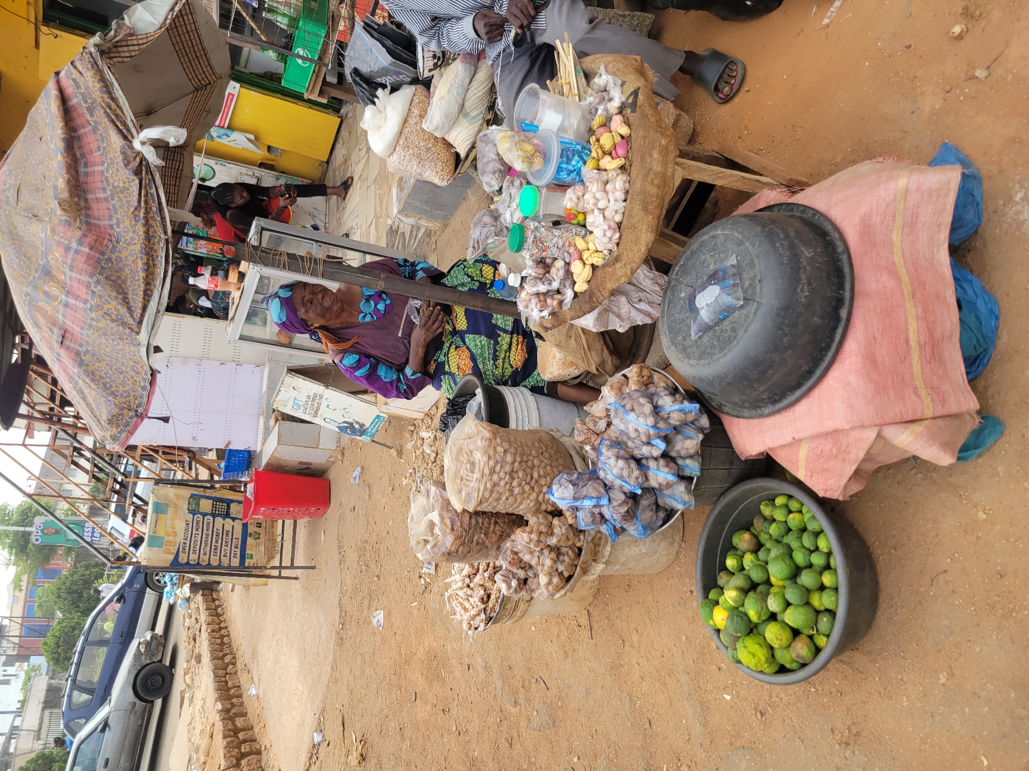The image depicts a bustling outdoor market scene in Jos, Nigeria, under bright daylight. The ground is unpaved, suggesting a roadside or informal market setting.
Several individuals are present, engaging in market activities. In the foreground, a woman wearing a vibrant purple and blue patterned dress and headscarf is seated, appearing to be a vendor. Beside her, a man in a striped shirt and grey trousers is also seated. Further back, another person in black is visible behind a counter.
The ground around the main stall is filled with a variety of goods for sale. These include numerous clear plastic bags and hessian sacks containing nuts or seeds (such as groundnuts or tiger nuts), piles of white garlic bulbs, yellowish root vegetables (likely ginger), and large black plastic bowls filled with small, green, lime-like fruits. Various small plastic containers holding candies or other items are also displayed. A makeshift canopy, fashioned from patterned fabric, provides shade over part of the stall.
In the background, a blue car is parked on what appears to be a street, further indicating the market's location along a thoroughfare. Visible signage includes a poster advertising financial services such as "OPEN ACCOUNT," "DEPOSIT MONEY," "WITHDRAW MONEY," "SEND MONEY," "BUY AIRTIME," and "PAY BILLS," alongside a mention of "GIFT ADATAPLAN TO YOUR FRIENDS AND RECEIVE CASH" and "PRAPTION CENTRE." The scene captures the everyday commerce and energy of a local Nigerian market.