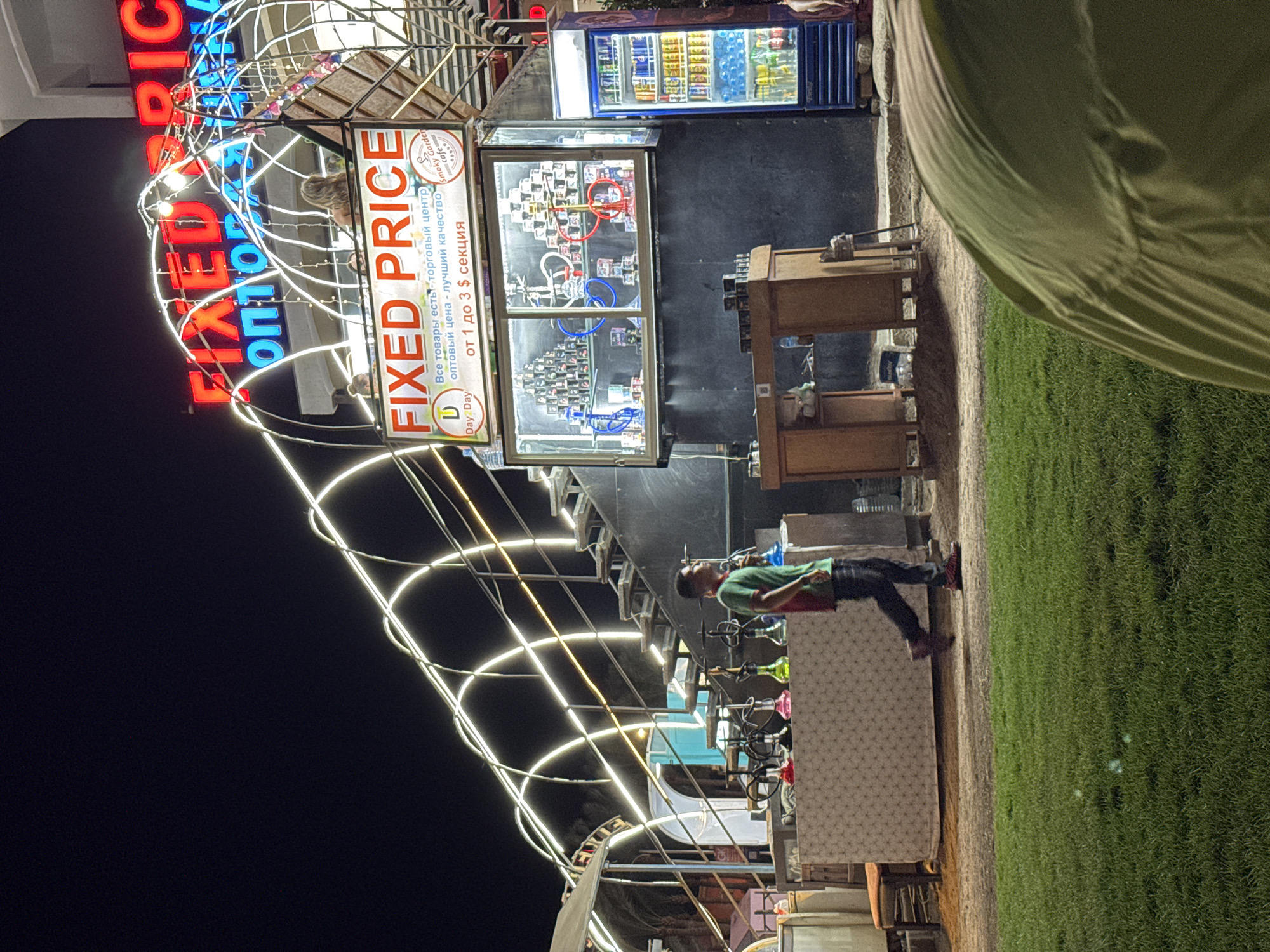 At night, on a paved surface in front of a grassy area, a young man wearing a green shirt and black pants walks past a display of hookahs. Behind him, a glass-fronted stall displays numerous boxes of what appear to be shisha tobacco, arranged in stacks. A refrigerator filled with beverages is to the right of the stall. Above the stall, a neon sign reads "FIXED PRICE" in red letters. Further up, a larger neon sign, partially obscured, also features red lettering. String lights curve over the scene, creating an arching effect. A few people are visible on an elevated platform behind the stall. The overall impression is of a marketplace or shop in a tourist area.