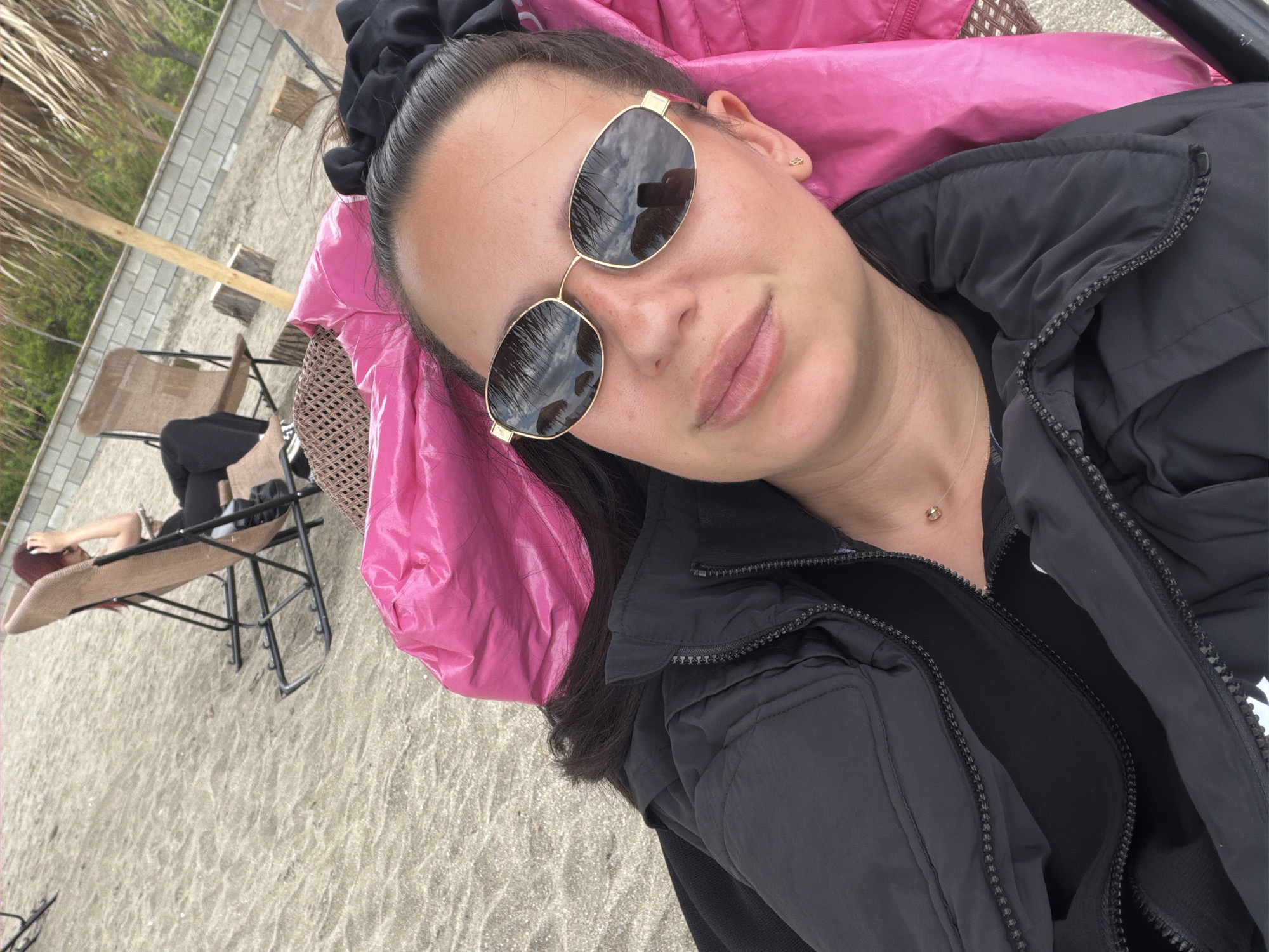 A woman wearing sunglasses and a black jacket is lying on a chair with a pink pillow. In the background, another person is reclining on a beach chair. The location is a beach in Burgas, Bulgaria.