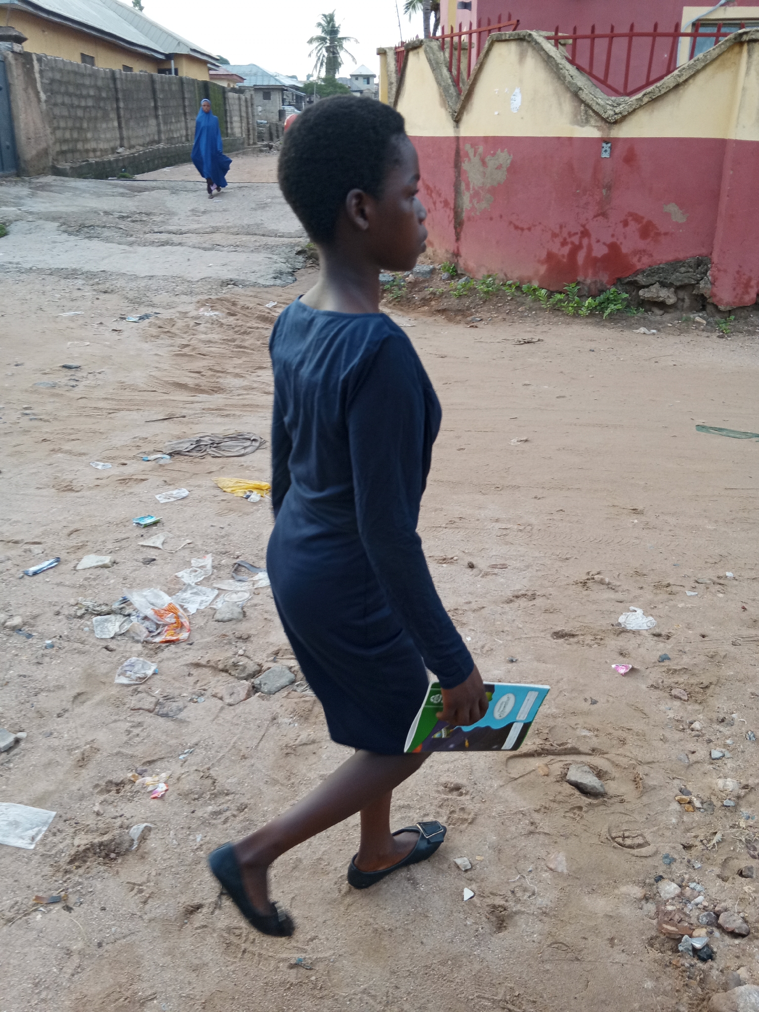 A young, dark-skinned woman with short, dark hair is walking on a dirt road littered with trash. She is wearing a long-sleeved, navy blue dress and black shoes. She is carrying a notebook in her right hand.
In the background, another person wearing a blue robe is walking away in the opposite direction. Buildings and a wall can be seen along the sides of the road. The location appears to be Zango-ndaji, Nigeria.
