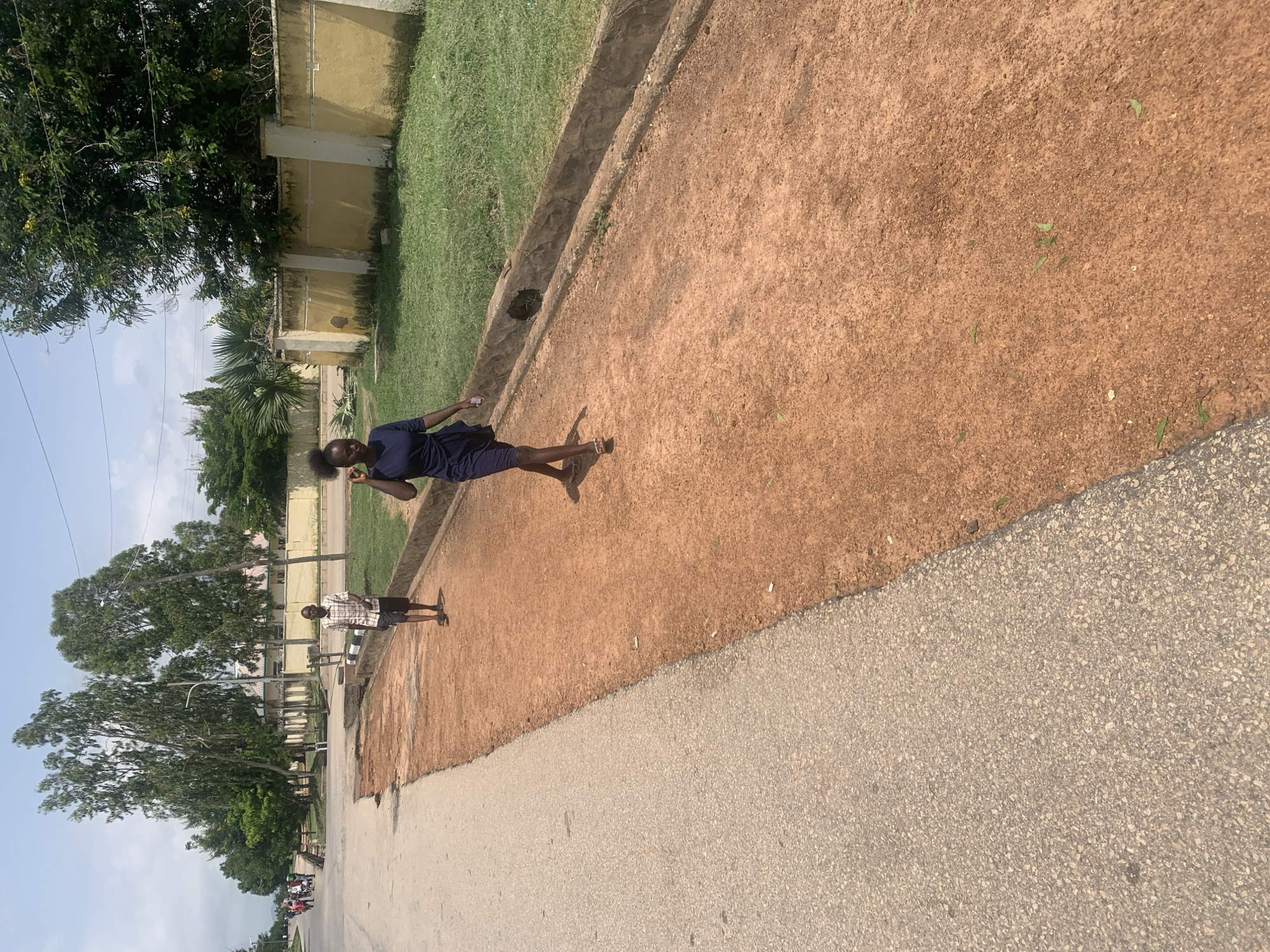 The image shows a person in a navy blue dress walking on a reddish-brown dirt path. The person is facing away from the camera, and their hair is in a ponytail. To the left of the path is a grassy area bordered by a concrete structure. Beyond the grass, a yellow building with a barbed wire fence on top is visible, along with trees and palm trees. The sky is blue with some white clouds.
Further down the path, another person is standing, wearing a plaid shirt and shorts. This person is also facing away from the camera. In the background, there are more trees and buildings. The ground is a mix of dirt and asphalt. The overall scene appears to be an outdoor area, possibly a school or park, given the presence of buildings, grassy areas, and trees. The lighting suggests it is daytime, with the sun casting shadows. The image is oriented vertically. No text is visible in the image. The location context provided is Jalingo, Nigeria.