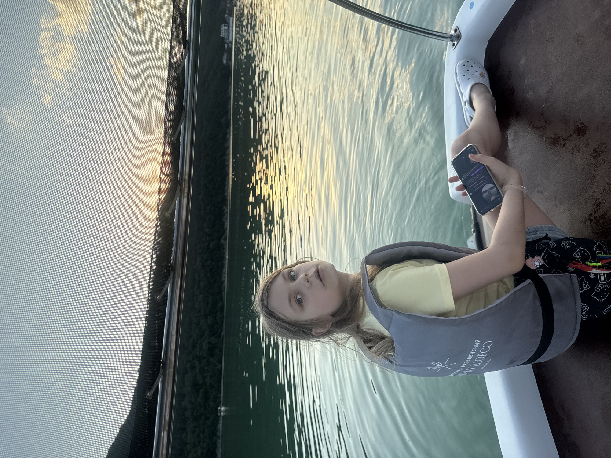In the city of Abrau-Dyurso, Russia, a young girl with blonde hair tied in a ponytail sits on a boat, looking over her shoulder towards the camera. She is wearing a yellow t-shirt and a grey life vest with white writing and a logo on the back. The girl holds a smartphone in her left hand. Her legs are extended, and she is wearing whiteCrocs. The boat is on a body of water, possibly a lake or reservoir, with calm waves reflecting the sunset. In the background, lush green hills line the opposite shore, and a few buildings are visible. The sky is a soft blue with hints of orange and yellow from the setting sun, partially obscured by a mesh screen or canopy above the boat.