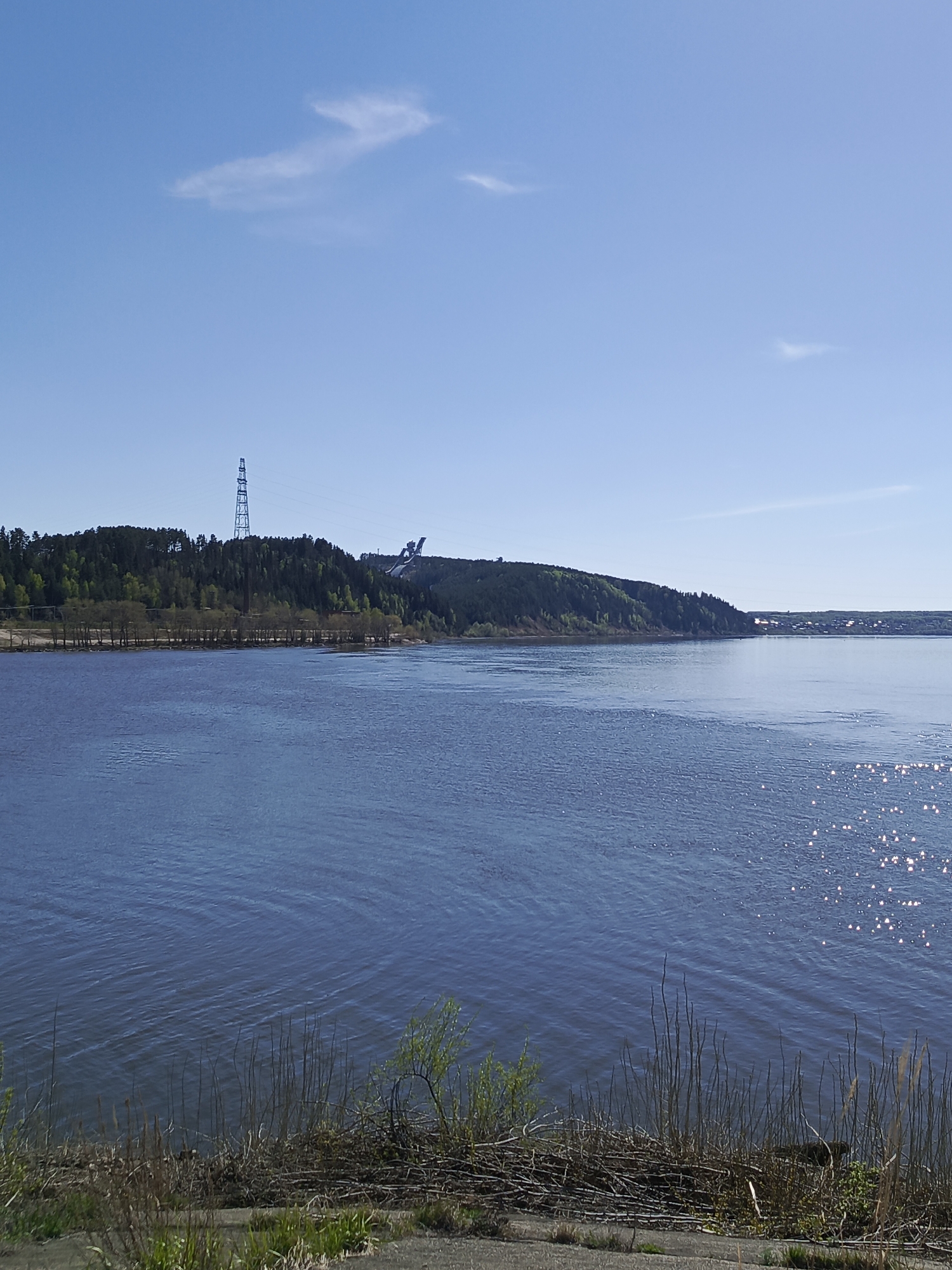 The image depicts a wide river under a clear, bright blue sky, with a wooded shoreline in the mid-ground. Sparkling sunlight reflects on the water's surface on the right side of the frame. On the left side of the river, the tree line is lower, with a few sparse trees visible near the water. Further inland from the left tree line, a prominent, tall electrical transmission tower is visible atop a hill. To the right of the transmission tower, on another hill, a large ski jump complex is visible against the clear sky. Reeds and some brushy vegetation occupy the foreground along the river's edge. A section of concrete is at the very bottom edge of the frame, suggesting a waterfront walkway or structure. The image is likely taken near the city of Chaikovsky, Russia, due to the presence of the ski jump, a well-known landmark of the city. The scene is serene and natural, with evidence of human infrastructure.