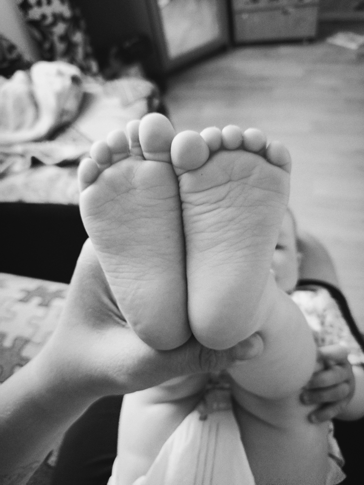 A close-up, black-and-white photograph shows a pair of infant feet being held up by an adult hand. The soles of the feet are facing the viewer. The toes are clearly visible and rounded. The hand holding the feet appears to be an adult's. Part of the baby's body is also visible. The background includes a patterned surface and a piece of furniture, possibly a bed or sofa. The location is not discernible.
