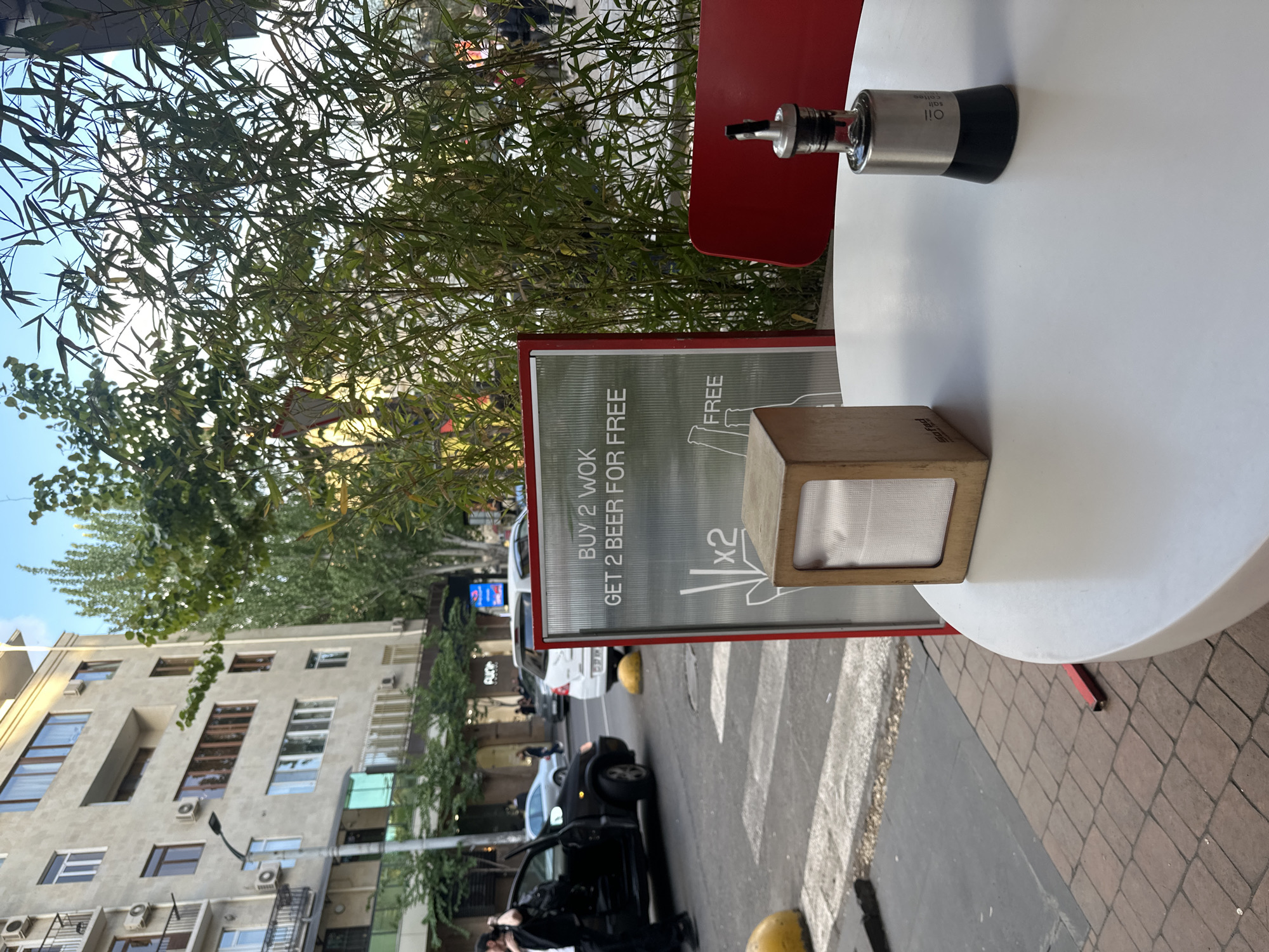 Here is a detailed description of the image:
**Scene Overview:**
The image captures a scene at an outdoor cafe in what appears to be a busy street in a city. The perspective is from a table with various objects and signs.
**Location:**
Although not explicitly stated in the image, the OCR-detected text "Safed" might indicate the brand, establishment, or location.
**Objects and Details:**
* A white round table is visible, with a metal container labeled "Oil Salt Coffee" on it.
* A wooden napkin holder with napkins is also on the table.
* A sign with red borders announces a promotional offer: "BUY 2 WOK GET 2 BEER FOR FREE x2"
* A lush green plant or tree is behind the table, providing a partial backdrop.
* The street scene includes a crosswalk, cars, and pedestrians. One individual is standing near a black car with the door open, possibly getting in.
* Buildings are visible in the background, some with multiple stories and windows.
**Environment:**
The setting is an urban environment with street-level businesses and residential buildings. The presence of trees and plants adds a touch of nature to the scene. The weather appears to be bright, possibly daytime.