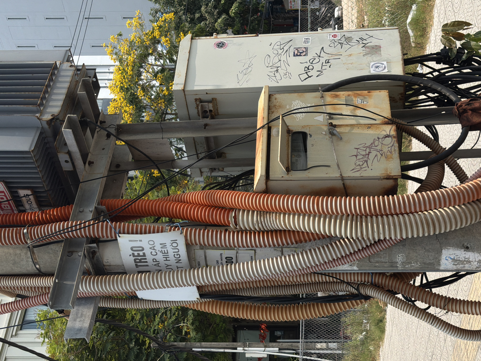 The media file captures a densely wired utility pole in Đà Nẵng, Vietnam, during the daytime under bright, clear weather. The central subject is a tall metal structure supporting various electrical components and a complex network of cables. On the left, a large, grey industrial transformer with cooling fins is visible. To its right, several smaller, weathered electrical boxes are mounted, showing signs of rust and covered in graffiti and stickers.
A prominent feature of the pole is the profusion of corrugated plastic conduits, predominantly in shades of orange, white, and brown, coiling around the pole and the electrical boxes. These conduits encase numerous black electrical cables, creating a tangled, yet organized, appearance. A white warning sign hangs amidst these conduits, partially obscured, with Vietnamese text.
In the background, a vibrant yellow flowering tree stands out against green foliage and parts of a building. The ground below features a paved sidewalk and some grassy areas behind a chain-link fence.
Visible text includes:
- On the warning sign: "TRÈO !" (Climb!), "ÁP CAO" (High Voltage), "NGUY HIỂM" (Danger), "CHẾT NGƯỜI" (Fatal/Deadly).
- Graffiti and tags on the electrical boxes: "NOT 38", "IBI", "RHOUS", and various illegible stylized writings.
- Numbers: "1909", "1930563", "252".
The scene depicts typical urban electrical infrastructure, highlighting the practical complexities of power distribution.