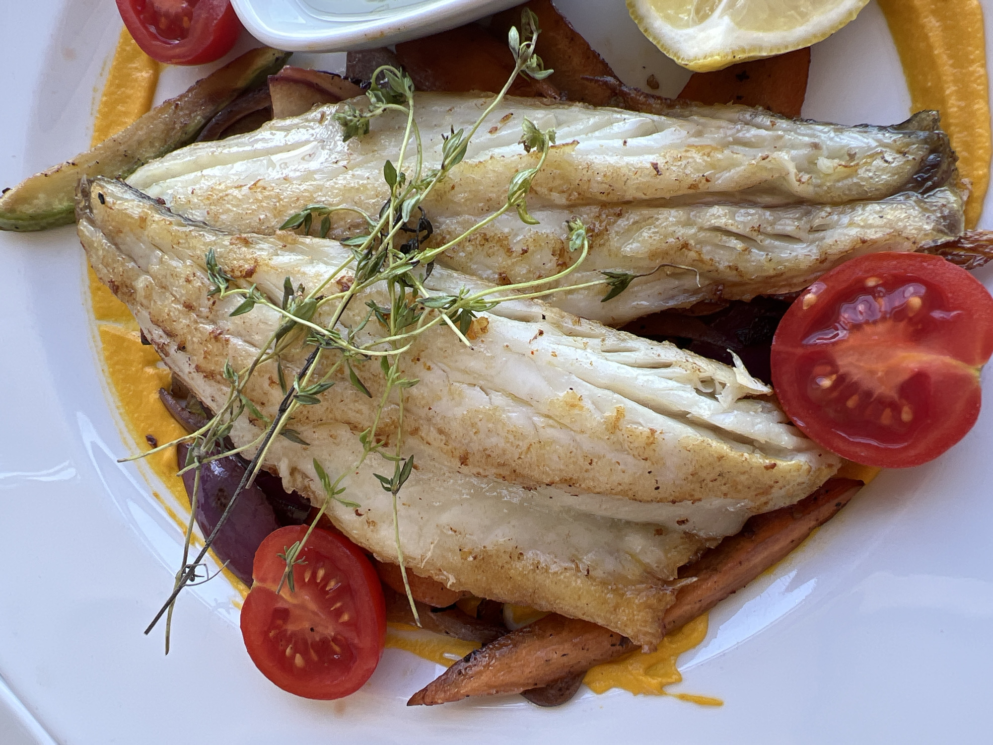 A close-up, overhead shot shows a prepared dish of pan-seared fish fillets served on a white plate. The fish, which appears to be white flaky fish, has a golden-brown crispy skin and is garnished with sprigs of fresh thyme. Beneath the fish, there's a bed of colorful vegetables, including sliced carrots, red onion, and possibly zucchini, all coated in a vibrant orange sauce. Halved cherry tomatoes and a lemon wedge are arranged around the plate. The food is artfully plated, suggesting it is from a restaurant in Kovačko Polje, Montenegro. The lighting is bright, highlighting the textures and colors of the food. There are no people visible in the image, and the focus is entirely on the appetizing meal.
