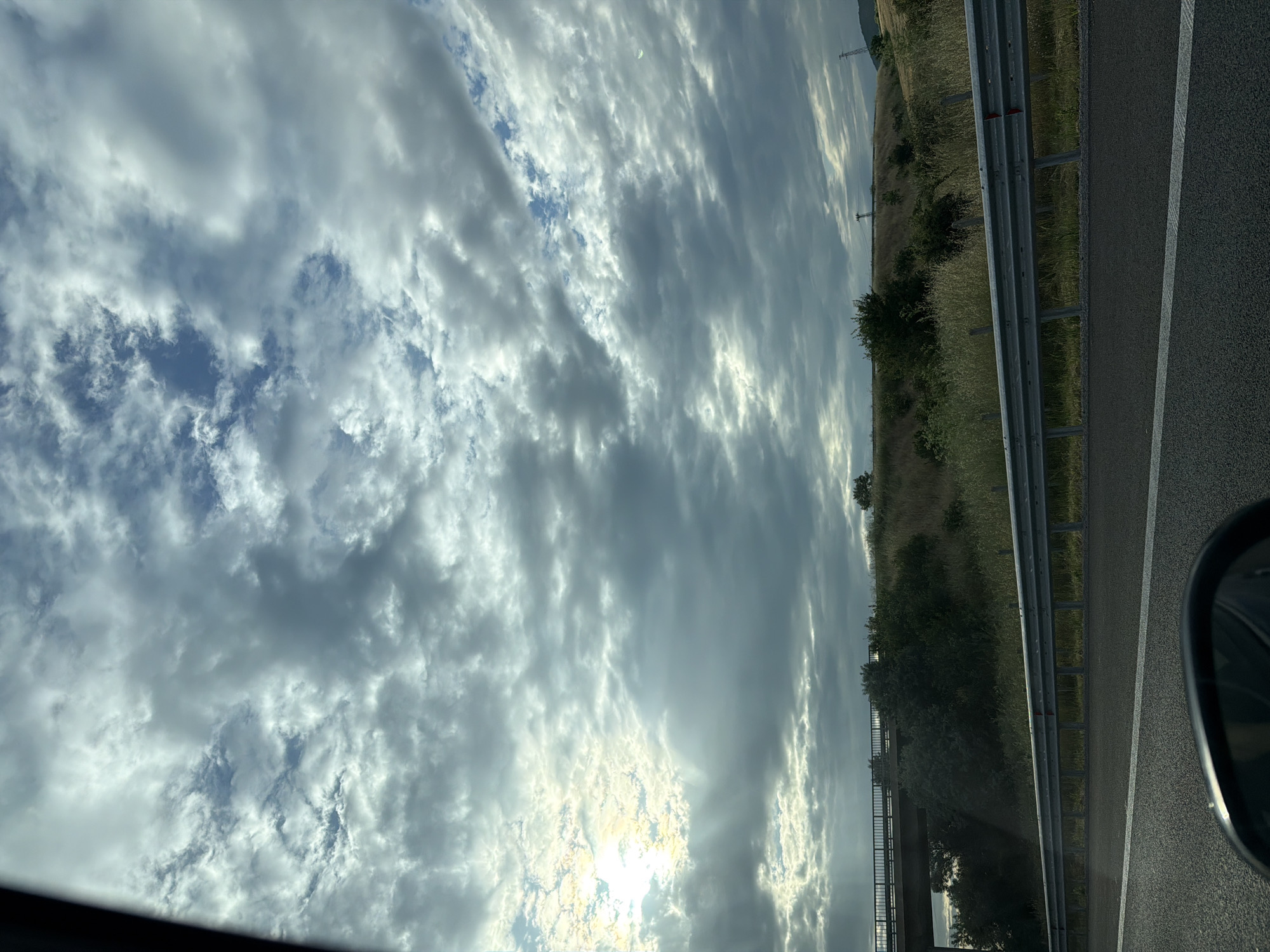 The image shows a view from a car on a highway near Polsko Padarevo, Bulgaria. The sky is filled with altocumulus clouds, with the sun shining through in the lower part of the frame. A metal guardrail is visible on the right side of the road, separating the highway from a grassy area with some bushes and small trees. A road is also visible behind the road's guardrail. A part of the car's side mirror is visible in the lower right corner. Power lines are also visible in the distance.