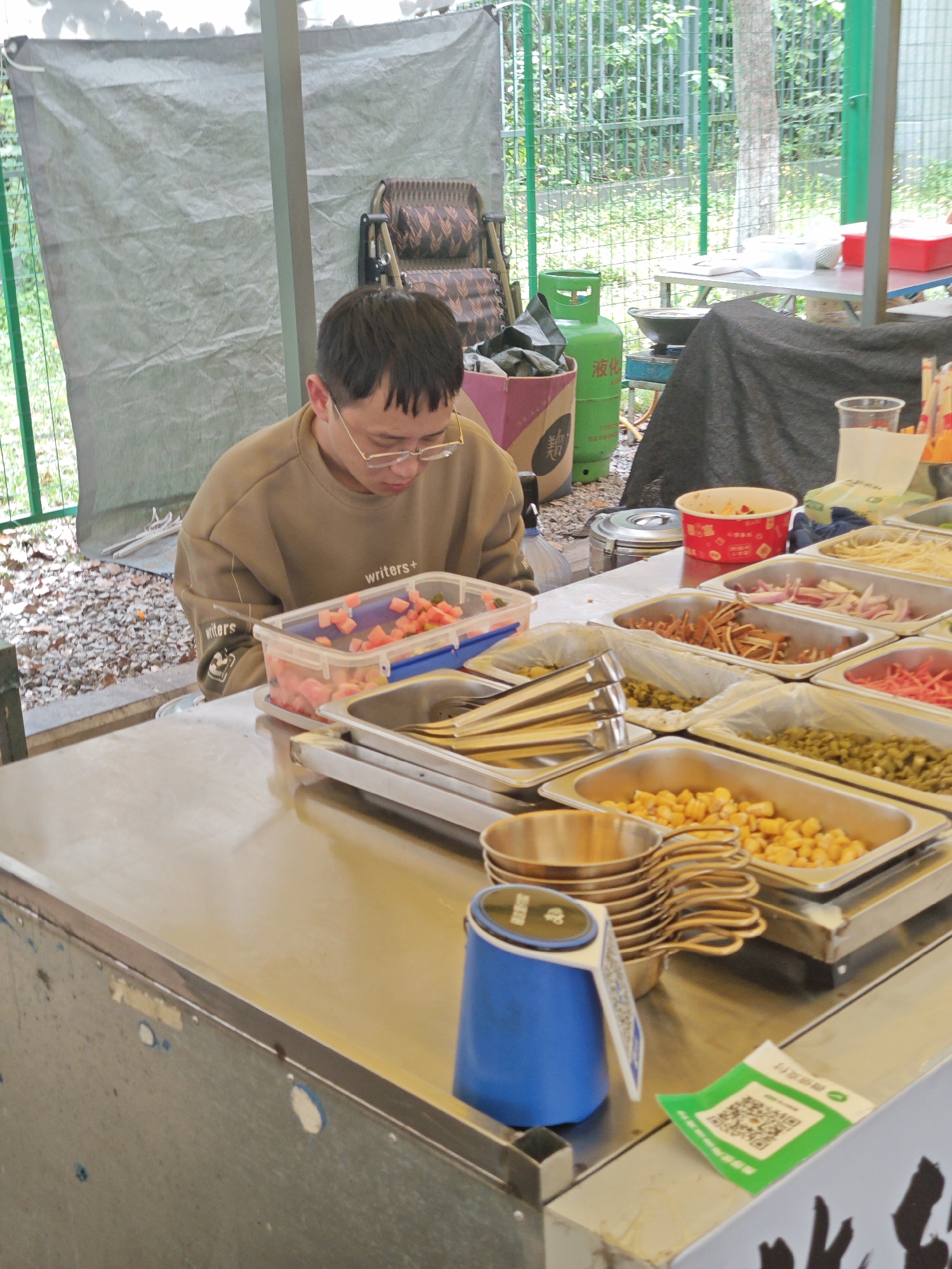 The image depicts a person wearing glasses and a light brown sweatshirt with "writers+" printed on it, standing behind a stainless steel counter. The counter holds several metal trays filled with various food items, including what appears to be chopped watermelon, noodles, red onions, some type of meat, and corn. To the right of the food trays, there is a stack of small metal bowls and a blue object with a white sticker. A green QR code is also visible on the lower right corner of the counter. In the background, there is a green propane tank and a green fence. The image was captured in the city of Shapingba District, China.