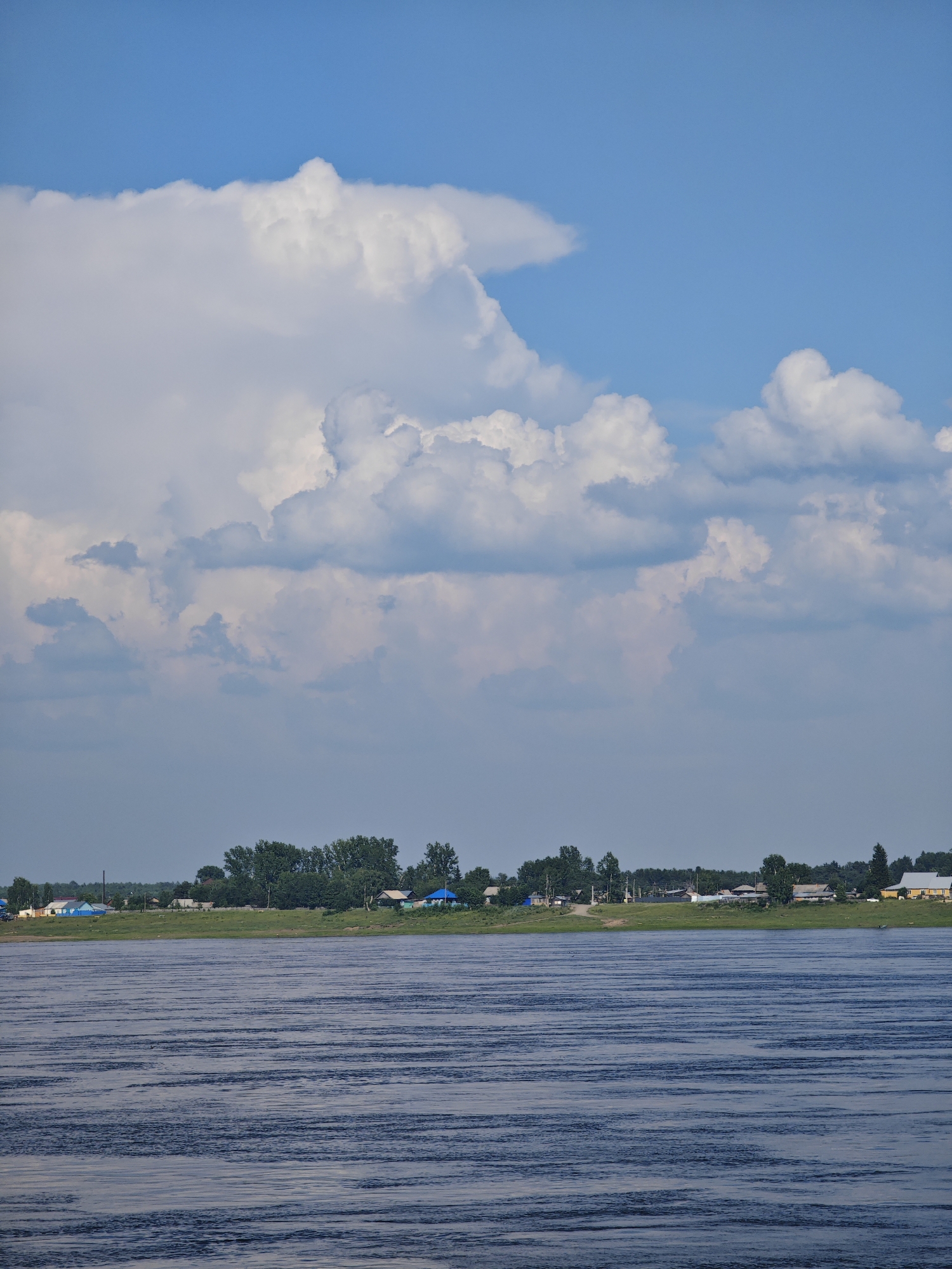 The image depicts a wide river in the foreground, with gentle ripples across its surface. In the middle ground, a green embankment is lined with trees and a village. The village consists of several small, scattered houses, some with brightly colored roofs, and appears to be situated in Verkhnepashino, Russia. The background is dominated by a vast blue sky with large, fluffy white clouds. The overall scene is serene and natural, suggesting a peaceful rural setting.