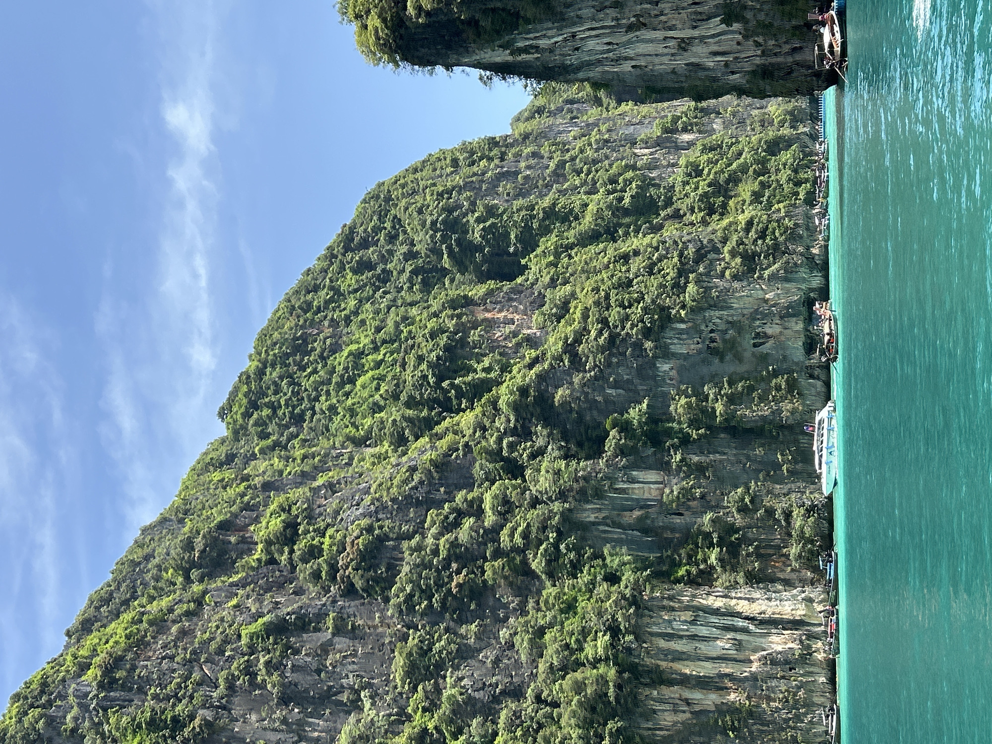 The image depicts a serene scene in Ao Nang, Thailand, showcasing the stunning limestone cliffs characteristic of the region, teeming with lush green vegetation. The turquoise waters of the Andaman Sea gently lap against the base of these majestic cliffs, reflecting the clear blue sky dotted with wispy white clouds. Several longtail boats and a speed boat are anchored in the water, some appearing to be carrying passengers, while others are idle. The overall impression is one of natural beauty and tranquility, a popular tourist destination.