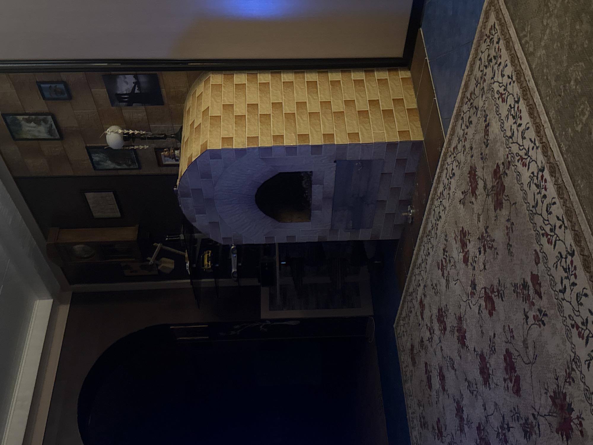 The image depicts an interior scene, possibly a living room, with a large floral patterned rug in the foreground. To the right, there is a brick-tiled oven or fireplace with a dark opening. Behind the oven, a wall is decorated with several framed pictures and a lamp. To the left, there is a dark arched doorway leading to an unseen area. The overall lighting suggests it is evening or indoors with artificial light. The image does not provide any specific clues to identify the location as being in Galinė, Lithuania, or any other specific city.