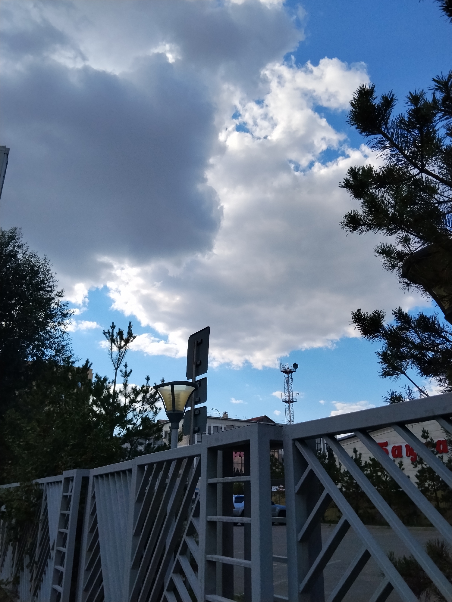 The image depicts a fence in front of a building with Cyrillic lettering. The fence is made of grey metal with a geometric pattern. Beyond the fence, trees are visible, and in the background, there is a telecommunications tower. The sky is blue with scattered clouds. This scene is located in Kokshetau, Kazakhstan.