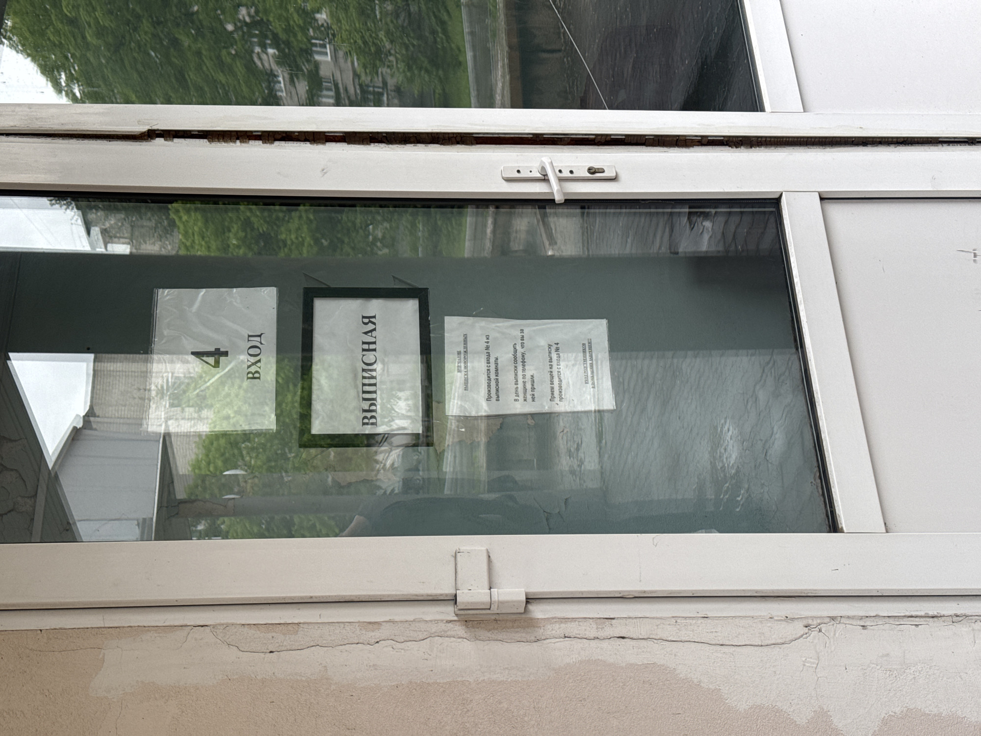 The image presents a close-up view of a window, likely part of an entrance to a building, possibly a maternity ward or hospital, located in Birobidzhan, Russia. The scene is illuminated by natural daylight, suggested by the reflections of green trees and another building on the glass, indicating a daytime setting.
Several informational signs, written in Russian, are taped to the inside of the window. On the left, a sign clearly indicates "4 ВХОД" (4 ENTRANCE). A central, larger sign reads "ВЫПИСНАЯ" (Maternity Discharge Room), with smaller text below it clarifying "ВЫПИСКА НОВОРОЖДЕННЫХ" (Discharge of Newborns). Further instructions are provided on a third sign: "Производится с входа № 4 из выписной комнаты" (Takes place from entrance № 4 from the discharge room). It advises, "В день выписки сообщить женщине по телефону, что вы за ней пришли" (On the day of discharge, inform the woman by phone that you have come for her). The sign also specifies, "Прием вещей на выписку производится с входа № 4" (Reception of items for discharge takes place from entrance № 4). A significant restriction is stated: "ВХОД РОДСТВЕННИКОВ В ПОМЕЩЕНИЕ ЗАПРЕЩЕН" (ENTRY OF RELATIVES INTO THE PREMISES IS PROHIBITED).
The window features a white frame, with a visible white latch on the upper section and a hinge-like mechanism on the lower part. The upper part of the frame shows some wear, with a crack or damage revealing the material underneath. Below the window, a light-colored wall exhibits signs of aging with visible cracks and some peeling paint. No individuals are present in the image. The setting suggests a designated area for managing the discharge process of new mothers and their infants, with clear rules for visitors.