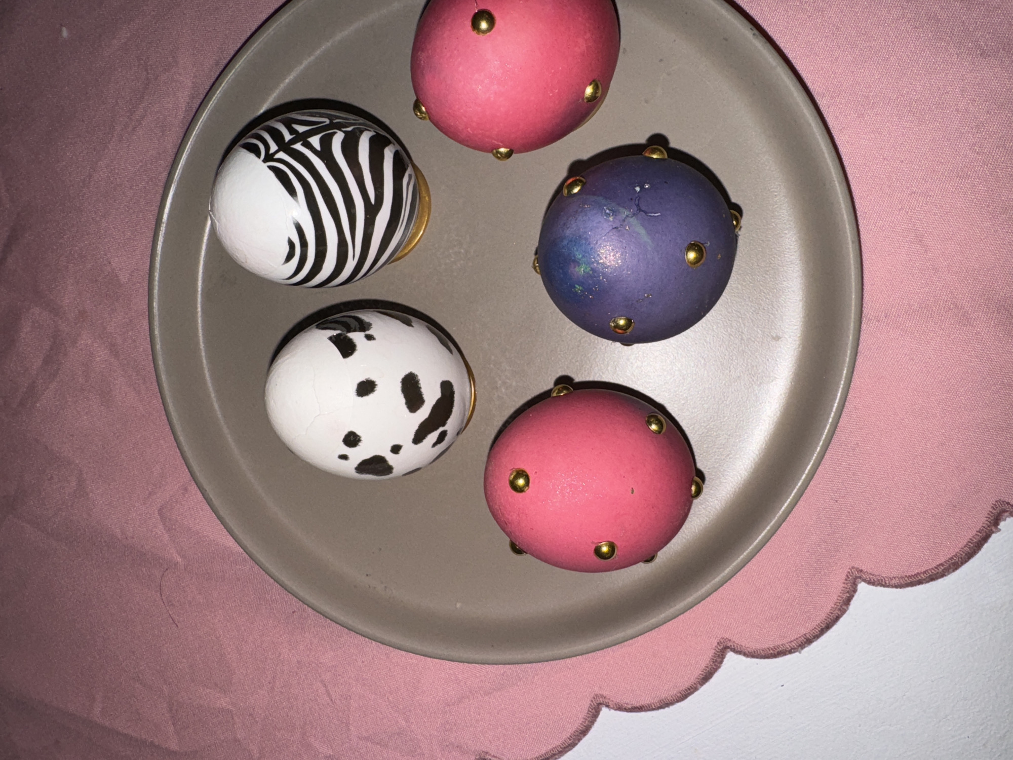 A top-down shot shows a gray plate with five decorated eggs on top of a pink cloth and a white surface. The eggs are arranged haphazardly on the plate. One egg has a zebra stripe pattern, and another has black Dalmatian spots. The other three eggs are solid colors: pink, red, and purple. Each of the solid-colored eggs is decorated with gold studs. The pink cloth is on the left side of the plate, and the white surface is on the right side. It's impossible to determine the location as Krasnoyarsk, Russia, with the given information.