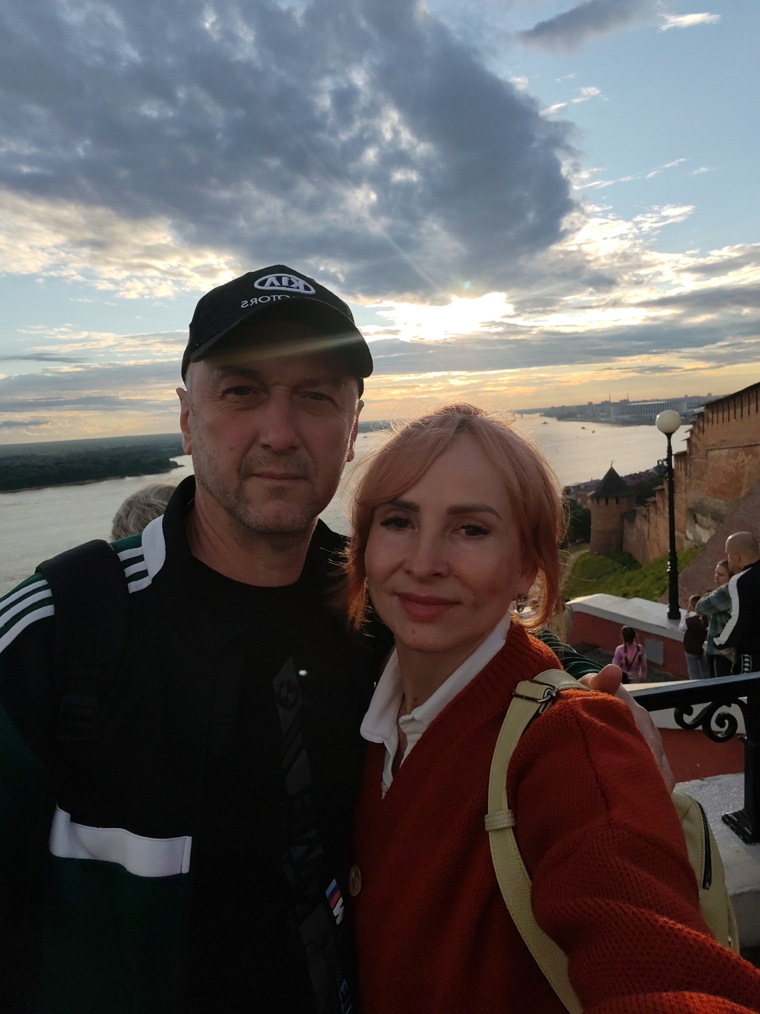 This media file features a selfie of a man and a woman in front of a scenic river and cityscape. The man, wearing a black baseball cap with a "KIA MOTORS" logo and a black jacket with white stripes, faces the camera with a neutral expression. The woman, with reddish-blonde hair, smiles gently, wearing a white shirt and an orange cardigan. She has a light-colored backpack slung over her shoulder.
The background reveals a wide river with a distant cityscape and buildings, including a historical fortress-like structure with brick walls and towers. The sky is dramatic, filled with clouds tinged with the warm hues of either sunrise or sunset, with the sun peeking through. The overall atmosphere suggests a pleasant outing or sightseeing. The visible text on the man's cap is "KIA MOTORS". The style of the architecture and the presence of the river suggest a location like Nizhny Novgorod, Russia, as hinted by the context. The time of day is either early morning or late evening, indicated by the lighting and sky.