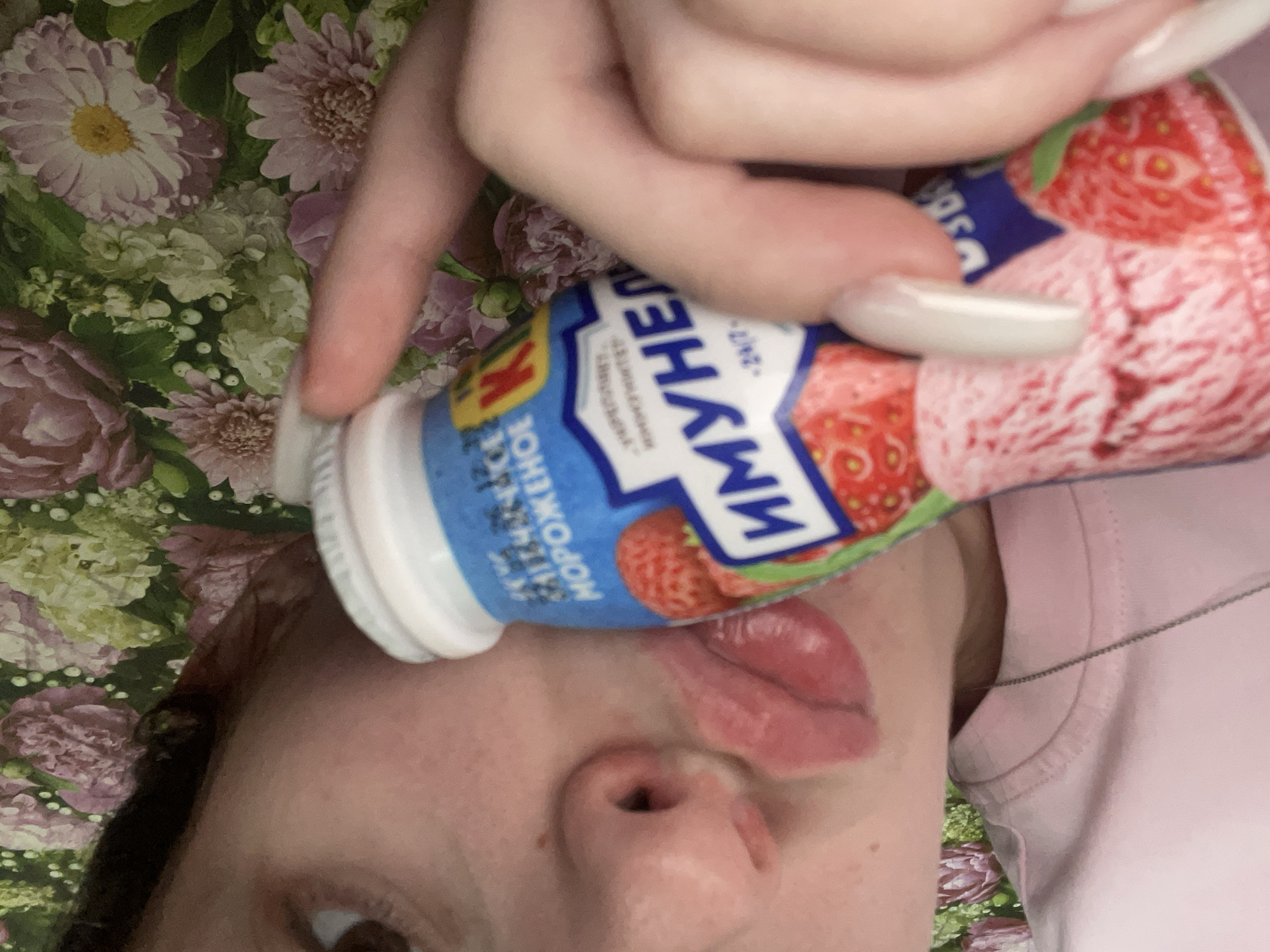 The image presents a close-up, rotated view of a person's face and hand, engaged with a bottled drink against a vibrant floral background. The primary subject is a person, with only a portion of their face visible, including their nose, mouth with an extended tongue, part of their chin, and one eye. A hand with long, light-colored nails grips a white plastic bottle. The person is wearing a pink garment, likely a t-shirt, and a thin silver chain is visible around their neck.
The bottle has a colorful label, predominantly blue and red, adorned with images of strawberries, indicating its flavor. Prominent Cyrillic text on the label reads "ИМУНЕЛЕ" (Immunelle), a recognized brand of fermented milk product. The label also displays a "К" symbol and the word "МОРОЖЕНОЕ" (Morozhenoe), meaning "ice cream," suggesting an ice cream-flavored variant. Small, partially visible text includes "24/7".
The activity captures the person interacting with the bottle, with their tongue playfully or eagerly extended towards the opening, possibly in the act of drinking or tasting. The background is a richly patterned fabric, possibly a blanket or bedspread, featuring an assortment of flowers in hues of purple, pink, white, and green. This setting suggests an indoor, relaxed environment. No specific time of day or weather can be determined. The image's angle is intimate and casual, consistent with the location context of Медведево, Russia, where the brand Immunelle is available.