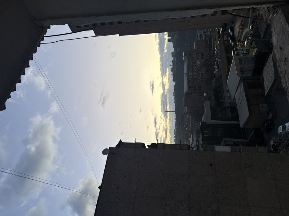 The image displays an elevated urban landscape in Yerevan, Armenia, captured during either dawn or dusk. The scene is framed by dark, unfinished architectural elements on the right, and the upper section of a dark, multi-story concrete building on the left, which features a white satellite dish on its roof and visible balconies on its right facade.
The sky occupies the upper half, transitioning from light blue directly overhead to a pale yellow-orange gradient at the horizon, indicative of low sun angle. Scattered white and light grey clouds are present, some illuminated from below. A thin, dark cable crosses the upper sky horizontally.
The midground reveals a dense urban environment comprised of various residential and commercial buildings, mostly appearing in shadow. Parked cars are visible on streets and in lots throughout the cityscape. A modern building with large glass windows and dark cladding is situated below the left foreground building. In the lower right midground, a fenced children's playground with colorful equipment, including slides and climbing structures, is visible.
The background consists of hilly terrain with additional structures and lights. A tall, slender industrial chimney or smokestack, characteristic of the Hrazdan Thermal Power Plant, prominently rises from the distant horizon slightly left of center. To the far right on the horizon, multiple construction cranes are discernible. No individuals or specific interactions are visible within the frame.