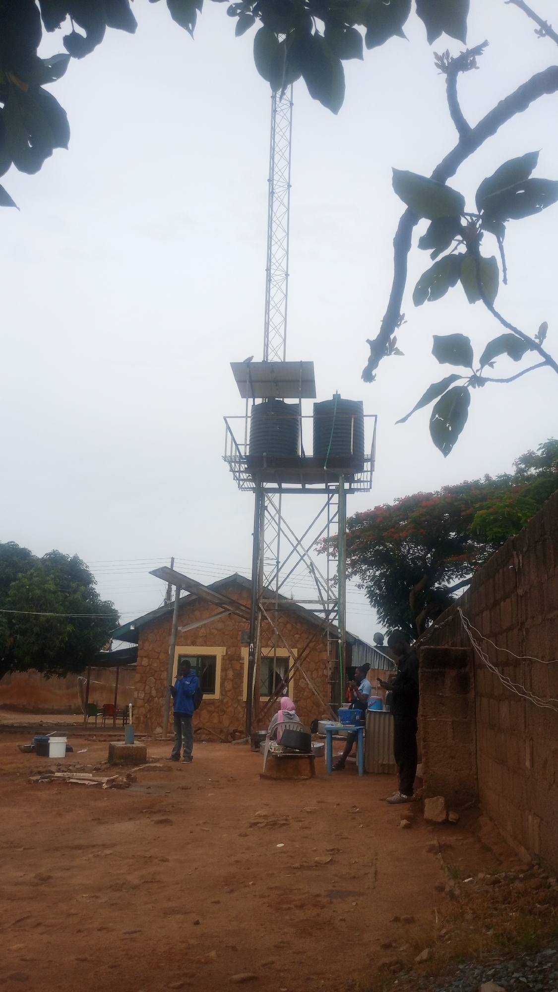 The image shows an outdoor scene in Bukuru, Nigeria. The foreground features a reddish-brown dirt ground. In the middle ground, a stone building stands with a corrugated metal roof. Adjacent to the building is a tall metal tower supporting two black water tanks and a solar panel. Several people are present: a man in a blue jacket is standing in front of the building, a woman wearing a pink headscarf is seated, and two individuals appear to be washing dishes at a blue table. Another person is standing near a brick wall to the right. A tree with green leaves is visible on the left, and a tree with red flowers is to the right. Overhead, leaves from trees partially obscure the top of the frame. The sky is overcast. Power lines are visible in the background. Miscellaneous objects are scattered around the dirt ground.