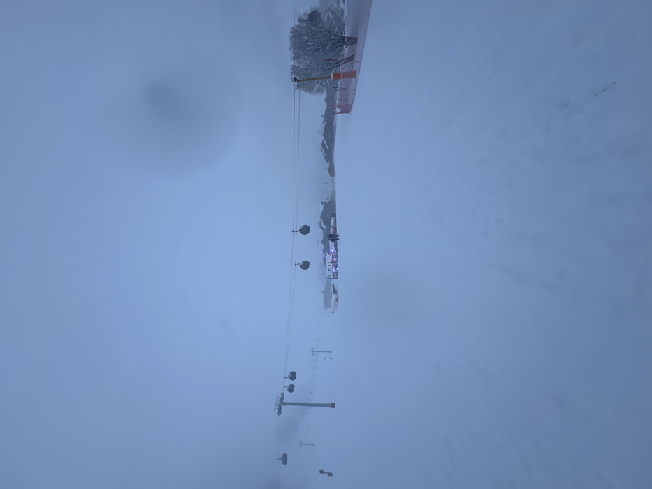 The image captures a snow-covered ski resort in Val-d'Isère, France, enveloped in extremely heavy snowfall or thick fog. The scene is dominated by muted tones of blue and white, indicating low light conditions and poor visibility.
A ski lift system is a prominent feature, with cables and several chairs suspended in the air, receding into the misty distance from the bottom-left towards the top-right. Multiple dark ski lift poles or towers are visible across the snow-covered terrain.
In the mid-ground, towards the top-right, a brightly illuminated building stands out against the white backdrop. This structure, possibly a lodge, restaurant, or ski station, has visible lights, suggesting it is either evening or night, or the intense weather conditions are obscuring daylight. A few snow-laden trees are clustered near the building, and some orange safety barriers or fences are also discernible. Several indistinct figures, likely people, can be seen near the lit building, possibly gathering or seeking shelter from the elements.
The ground is uniformly covered in a thick blanket of fresh snow, with no clear tracks or distinct features due to the flat light and pervasive haze. The heavy precipitation and low clouds merge the sky and snow, creating a diffuse, almost monochromatic landscape that conveys a sense of quietude and profound winter conditions at the resort. No visible text is present in the image.