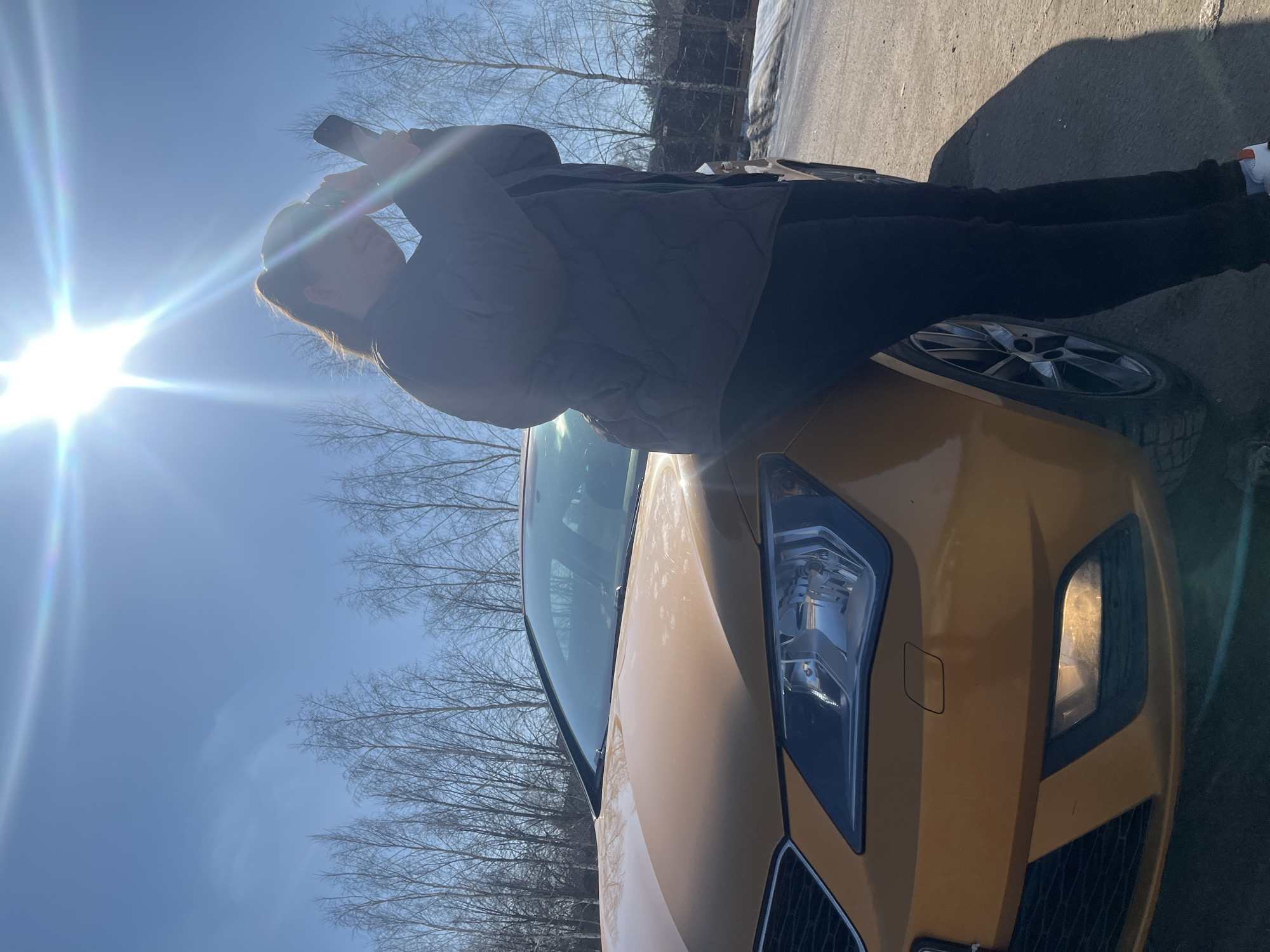A gold-colored passenger vehicle is parked on a dirt or gravel surface under a clear blue sky with strong overhead sunlight, causing lens flare. The front passenger side of the car, including its illuminated headlights, is visible. A female individual stands next to the vehicle, leaning slightly with her left arm resting on the hood. She is dressed in a dark quilted jacket and dark pants, with her hair pulled back. She holds a black smartphone device with both hands, raised to eye level, appearing to operate the device or shield her eyes from the sun. In the background, numerous bare-branched trees are visible, and distant patches of snow or ice are present on the ground. The specific location within Zubovo, Russia, is not discernable from the visual information provided.