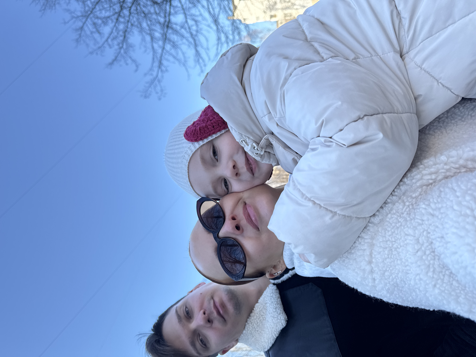 The image captures three people, a family consisting of a man, a woman, and a young child, posing for a selfie under a clear blue sky. The child, wearing a white winter hat with a pink heart detail and a puffy beige winter coat, is positioned above the woman, with their faces close together. The woman is wearing sunglasses and a white sweater. The man is below, slightly to the left. The background features bare tree branches and the corner of a building. The image was taken outdoors in daylight. The location is the city of Chaikovsky, Russia.
