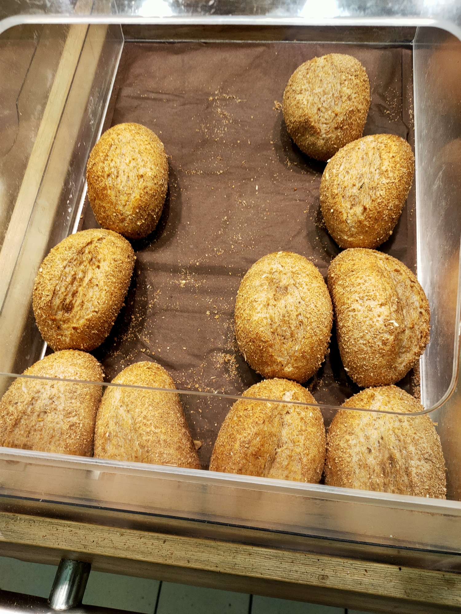 A display case in Kyiv, Ukraine, contains a variety of freshly baked bread rolls. The rolls are arranged on a brown paper liner within a clear plastic tray. The rolls are golden brown in color and appear to be coated in seeds or a similar grainy texture. Some of the rolls have a distinct scoring pattern on their surface. The display case is lit from above, casting a soft glow on the baked goods. The overall impression is one of a bakery or food market.