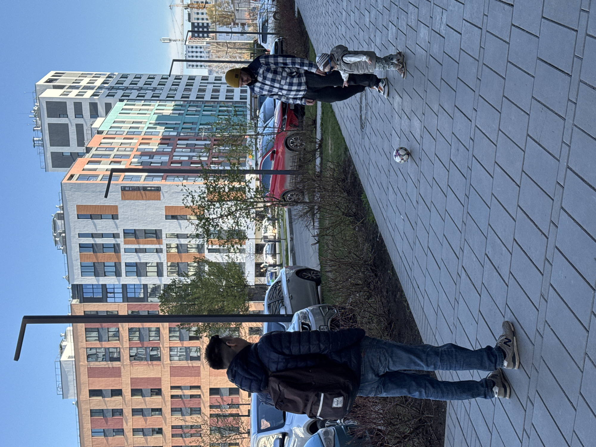 The image depicts an urban scene, likely a residential area, possibly in Izhevsk, Russia, given the style of architecture. The photo captures two men and a young child. One man is wearing a dark jacket and jeans with a backpack, standing on a paved walkway near a street with parked cars. In the distance, another man wearing a checkered shirt and a yellow beanie is walking alongside a young child, near a soccer ball. The environment includes modern apartment buildings, with visible cranes and construction in the background. Trees and shrubs line the walkway separating it from the street. The weather appears clear and sunny.